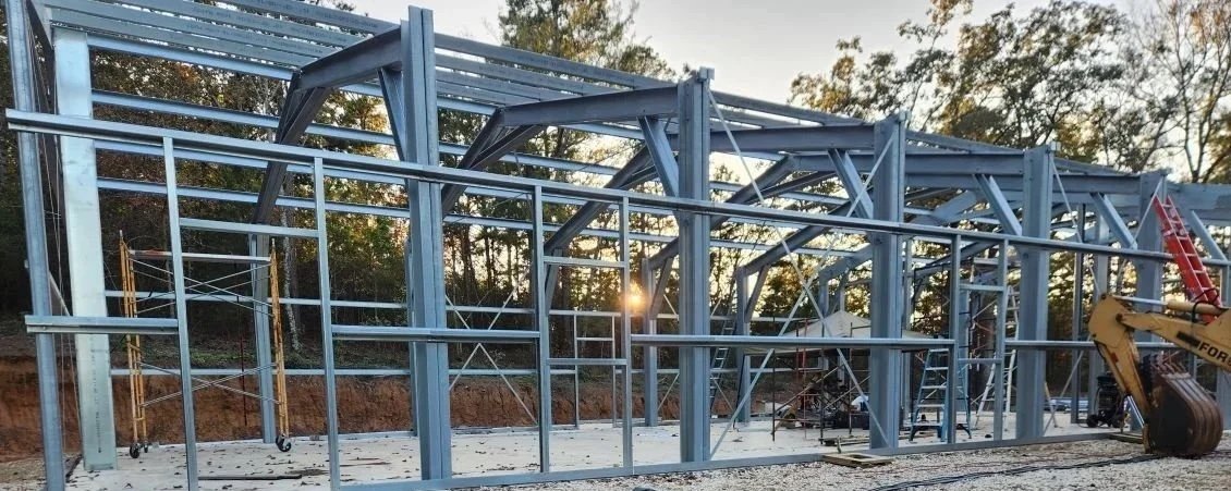 Steel framework of a building under construction during sunset, with trees in the background and construction equipment on site.