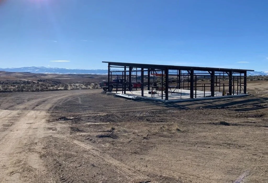 A partially constructed metal building frame in a flat, open, desert landscape with mountains in the distance and a clear blue sky.