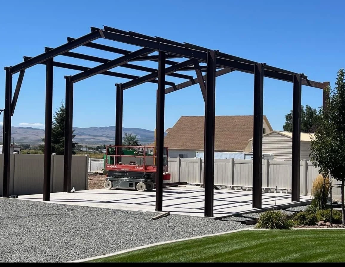 Under construction metal frame structure in backyard with gravel and grass, fence, and mountains in background.