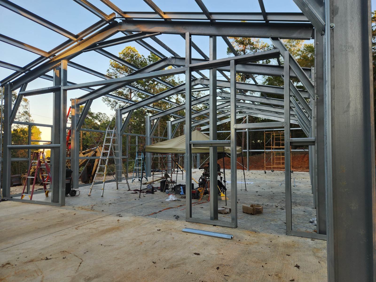 Metal framing of a building under construction with tools, ladders, and equipment on the ground inside the structure, and trees in the background.