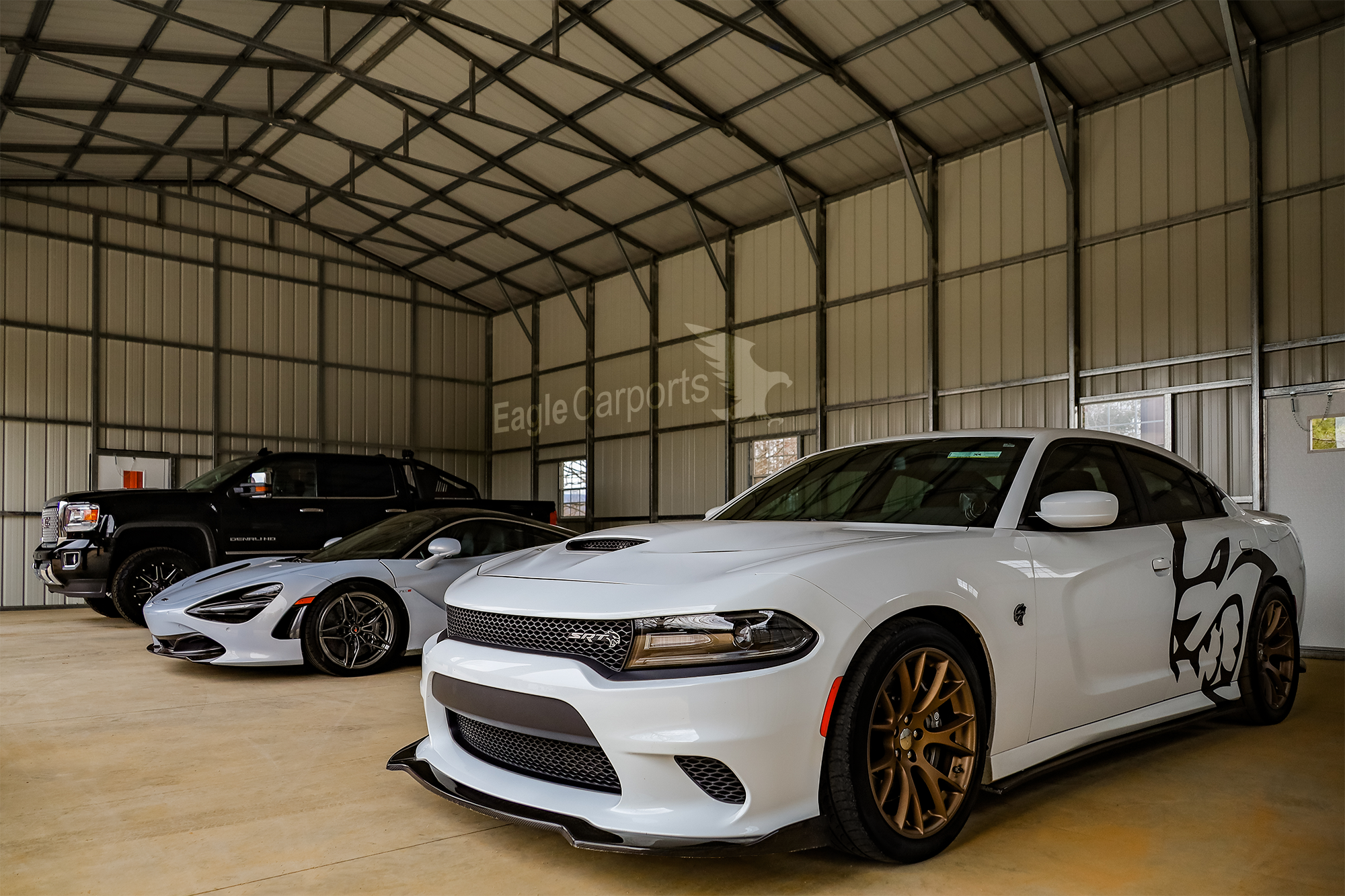 Inside a metal garage with three high-end vehicles: a white Dodge Charger with black and gold wheels and black graphic on the side, a silver sports car with black wheels, and a black pickup truck.