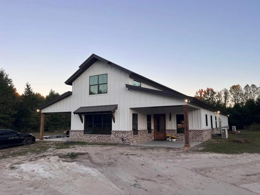 Newly built modern farmhouse with white vertical siding, black roof, front porch, brick foundation, and surrounding yard, under a clear sky.