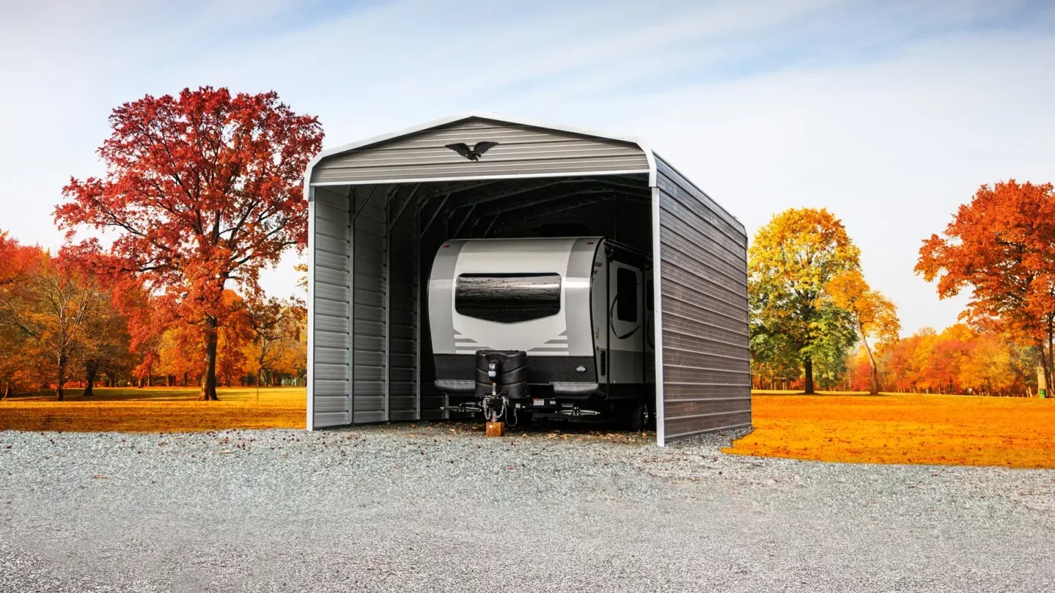 A white travel trailer parked inside a metal storage shed with a curved roof during autumn, with orange and yellow trees in the background and a gravel ground.