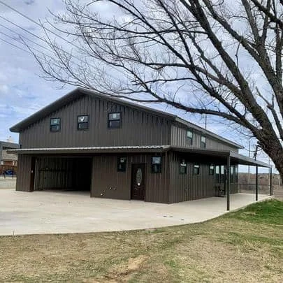 A two-story dark brown building with a sloped roof, large windows, and a covered patio area, with a large leafless tree in front and a grassy lawn in the foreground.