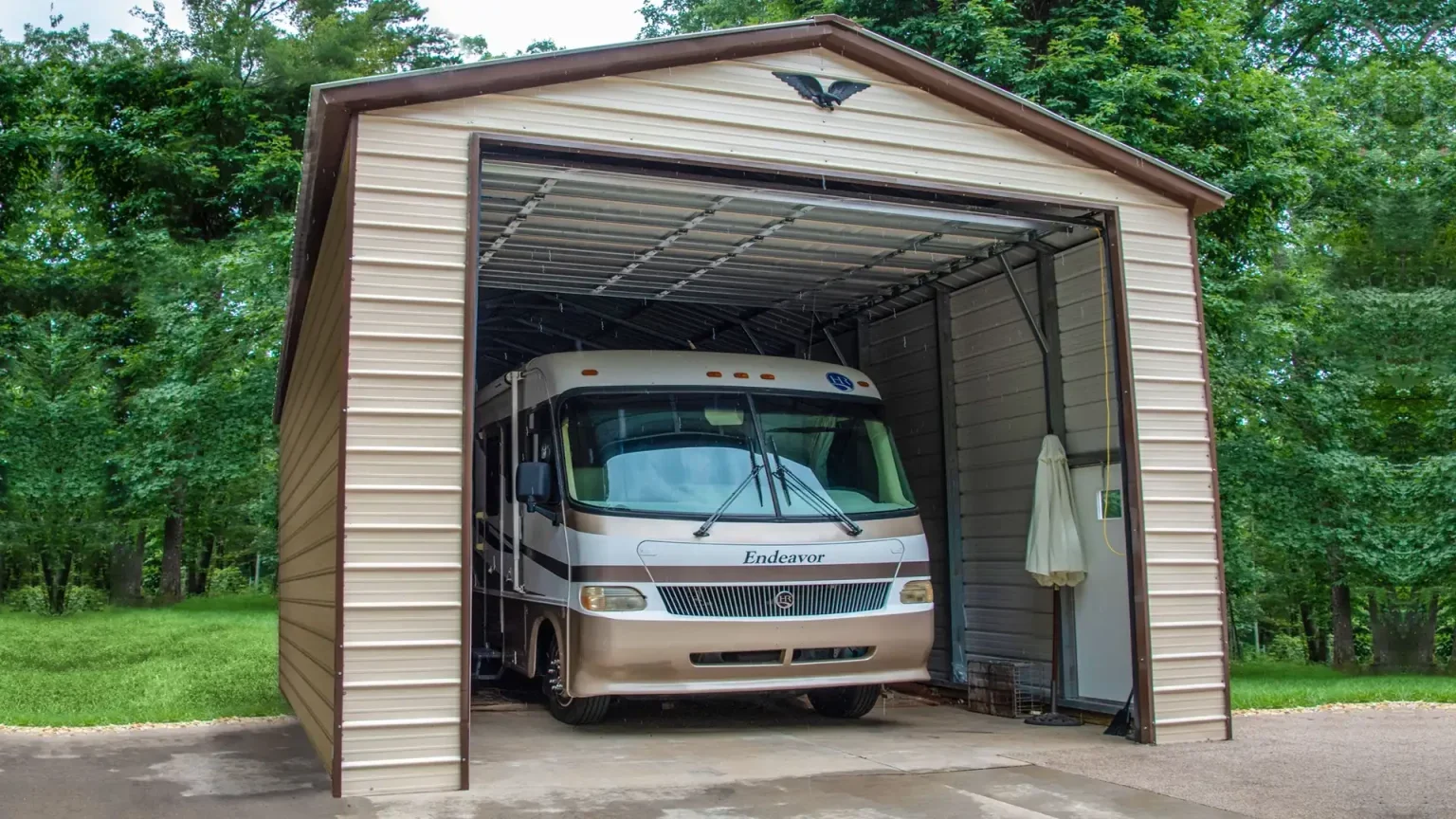 A beige and white RV parked inside a metal shed with an open front, surrounded by green trees and grass.