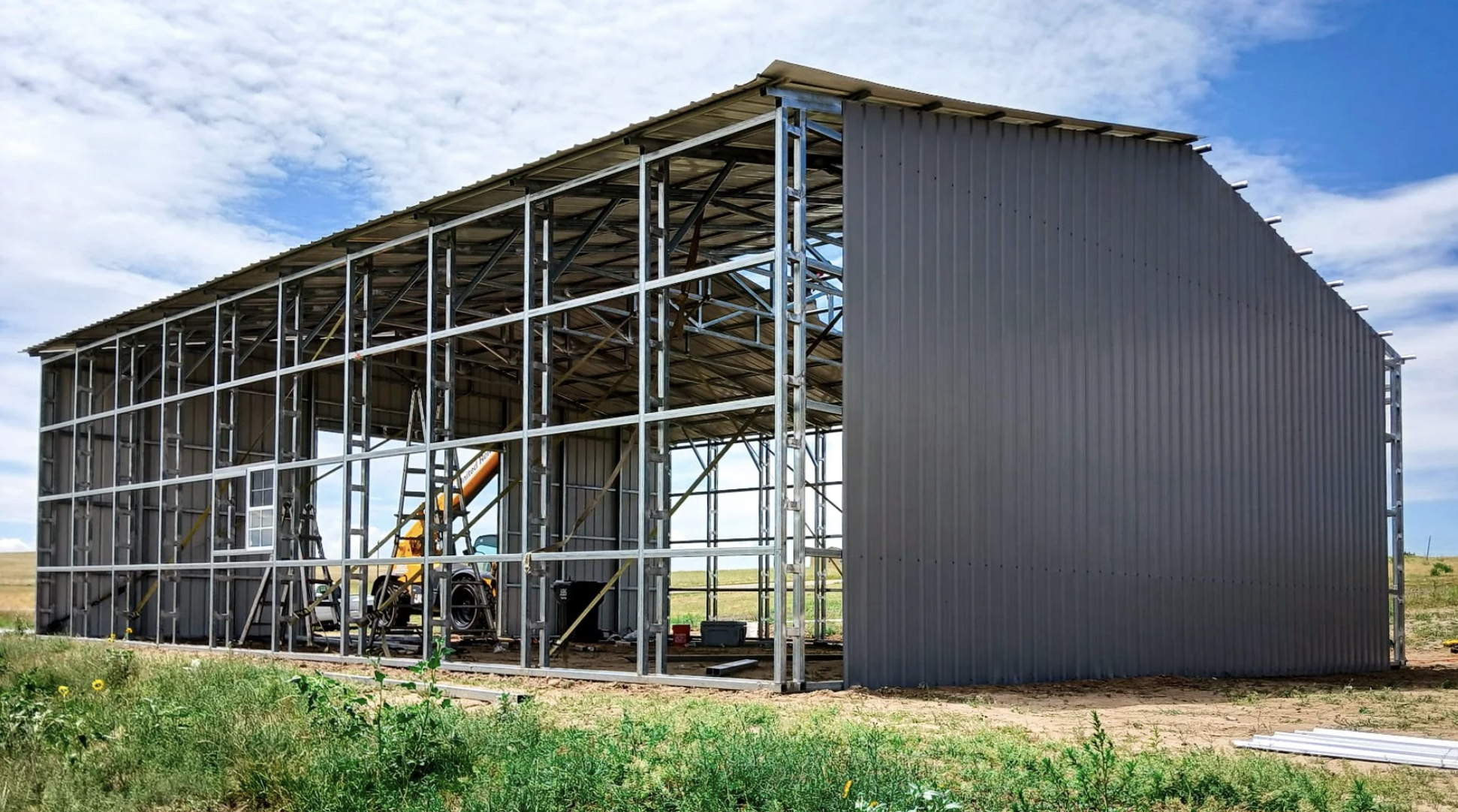 Construction of a large metal building with a steel frame and corrugated metal walls, partially built outdoors on a grassy field with a cloudy sky.