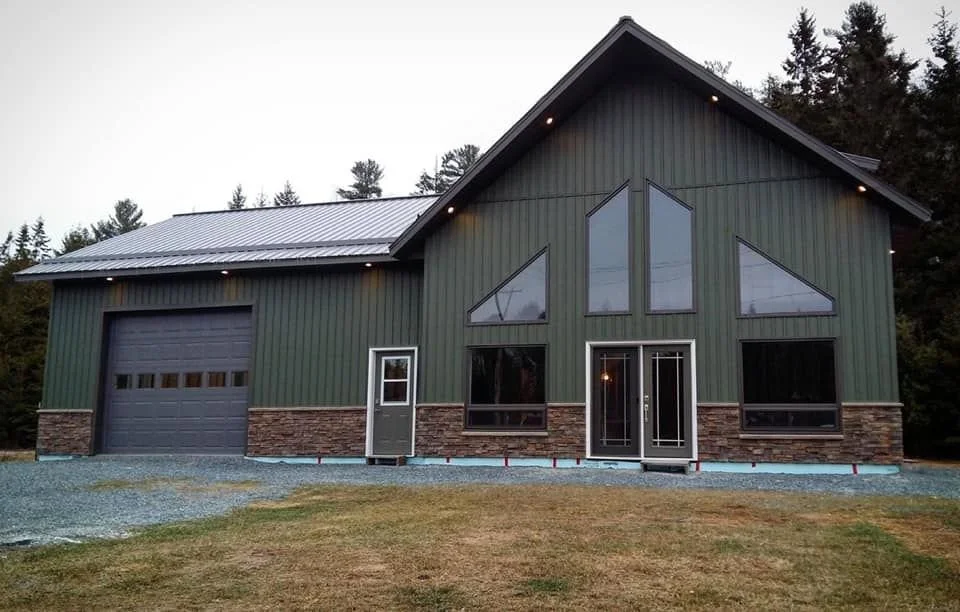 Modern two-story house with green vertical siding and stone accents, large front windows, a garage, and a front door, surrounded by trees.
