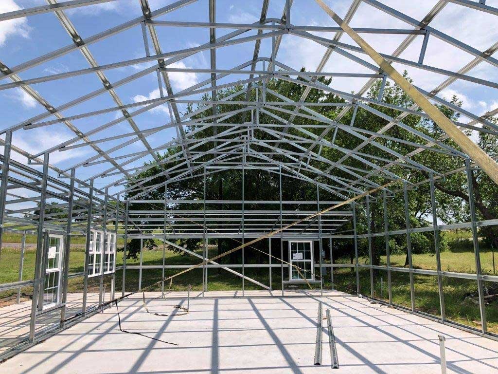 The image shows the metal frame of a greenhouse under construction with open windows and a concrete floor, surrounded by trees and grass.