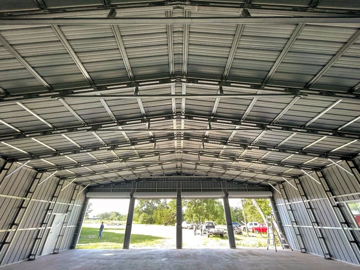 Interior of a metal garage or storage building with large open doors, showing outdoor parking lot and trees outside.