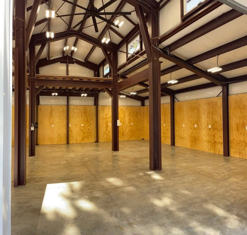 An empty spacious interior with concrete flooring, wooden wall panels, brown metal beams, and a high ceiling with multiple ceiling fans and lights.
