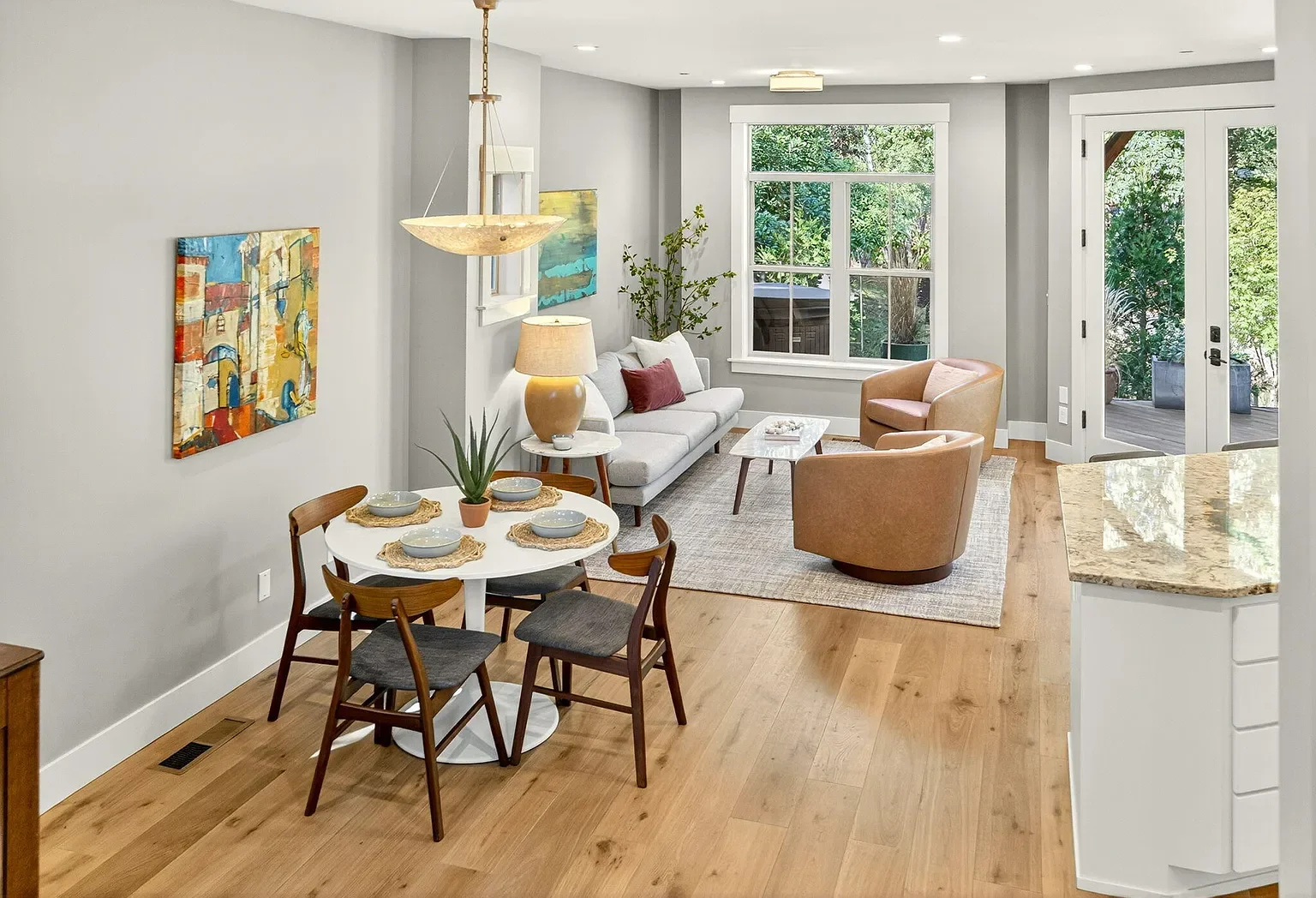Bright living room with light gray walls, hardwood floors, a white sofa with red pillows, two tan chairs, a coffee table, a window, and a door leading outside. There is a dining table with four chairs, artwork on the wall, and a lamp on a side table.