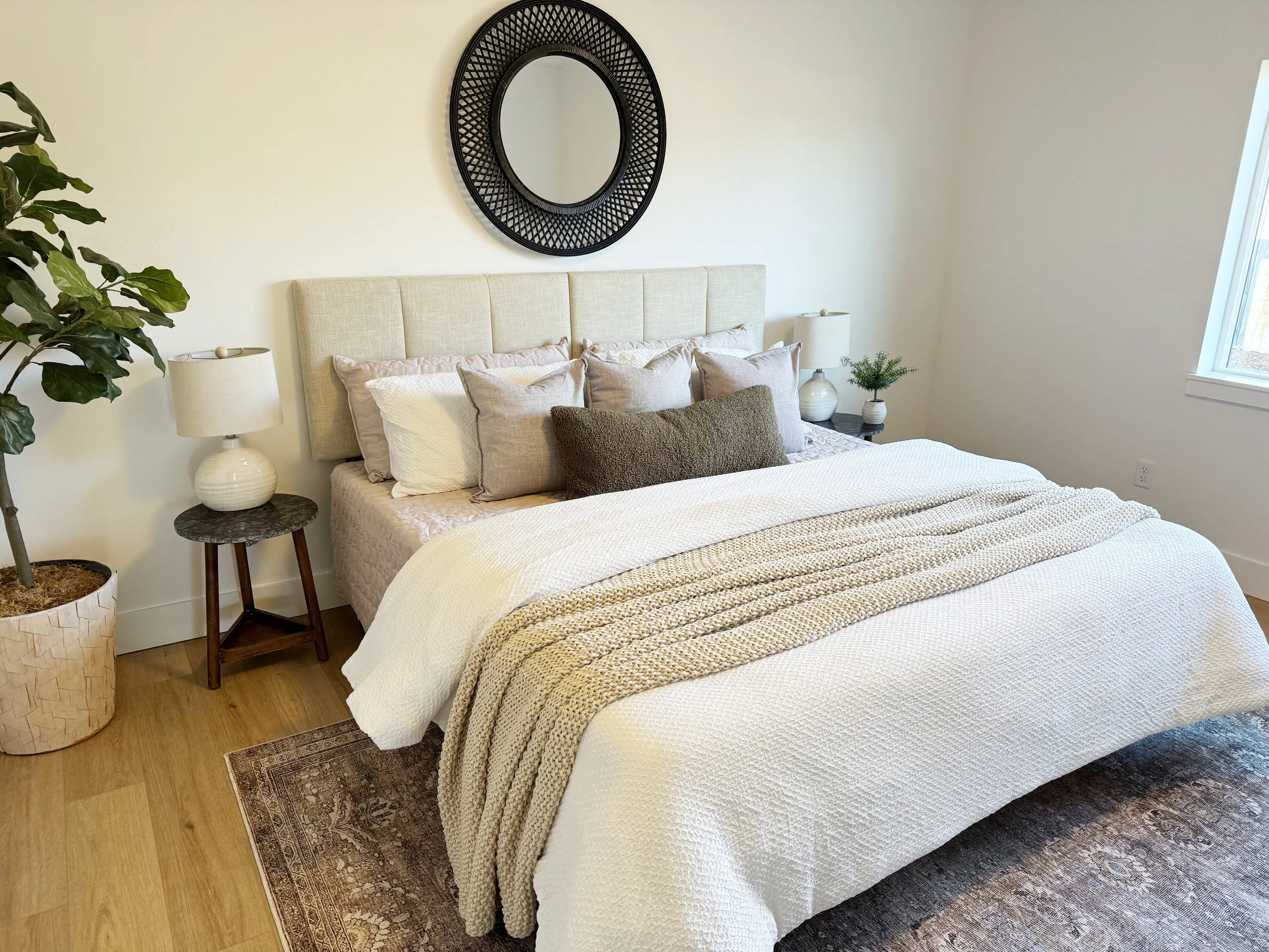 A neatly made bed with a variety of beige, white, gray, and brown pillows, a cream quilted bedspread, and a textured beige throw blanket. The bed has a beige upholstered headboard. Flanking the bed are two dark wood nightstands, each with a white cer