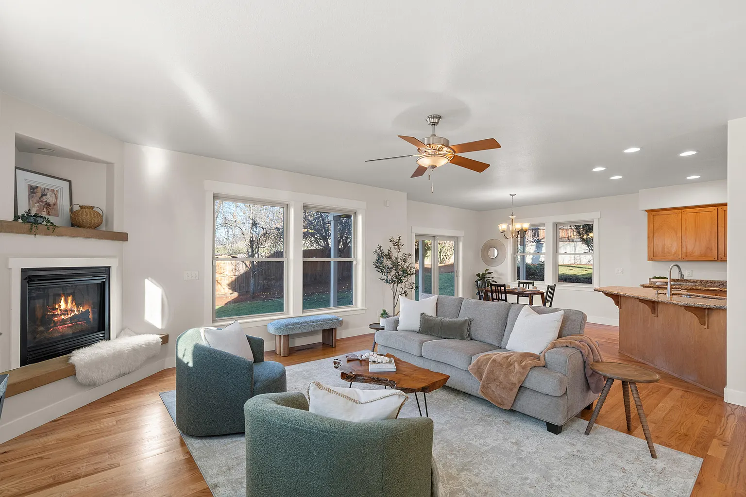 Open-concept living room with a gray sofa, green armchairs, a wooden coffee table, fireplace, large windows, and a dining area with a round table and chandelier, all with hardwood floors.