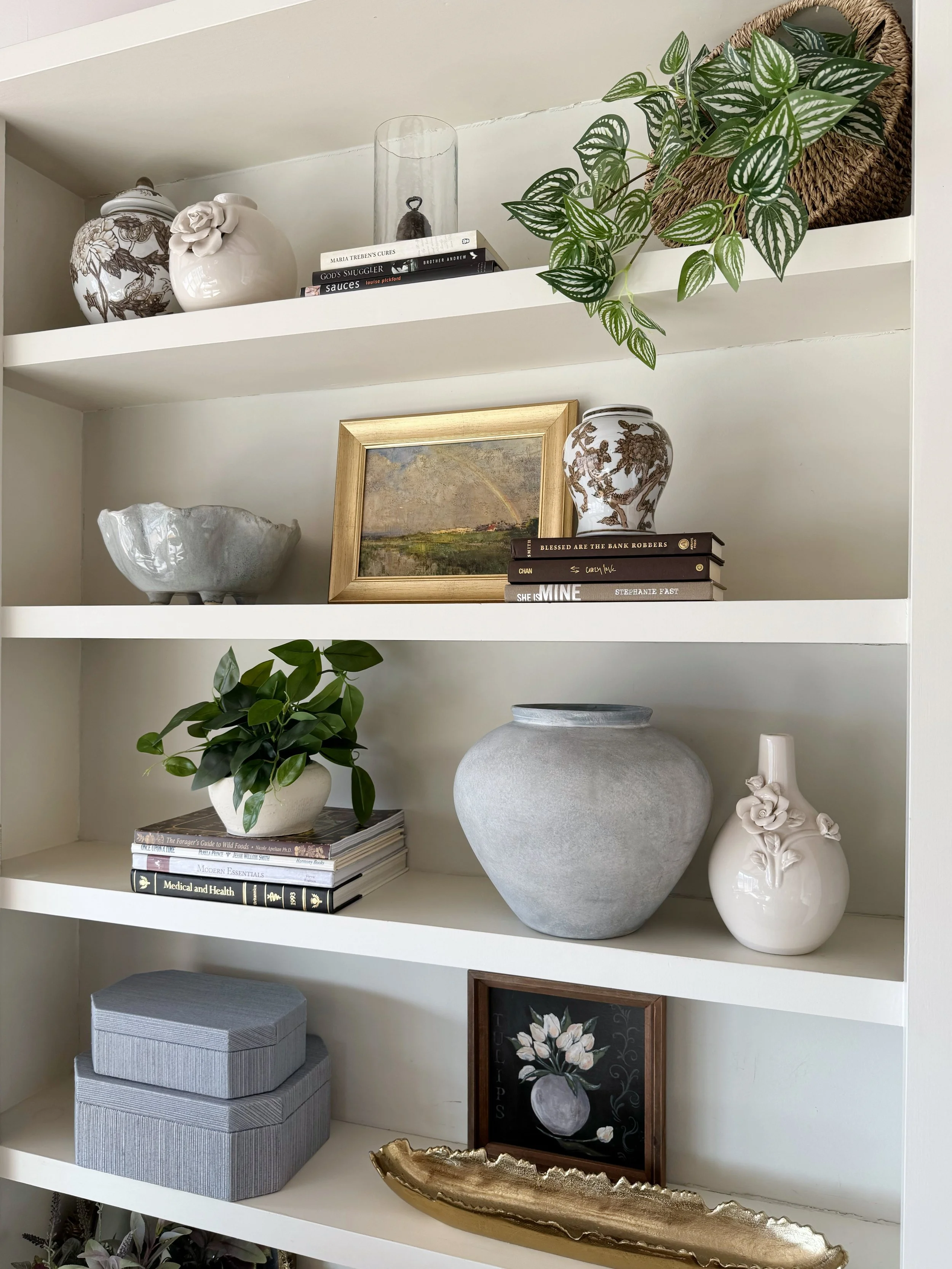White bookshelf with decorative vases, books, a framed painting, a potted plant, a gray decorative bowl, a large gray vase, a white ceramic vase with flowers, gray storage boxes, a framed floral artwork, and a gold decorative tray.