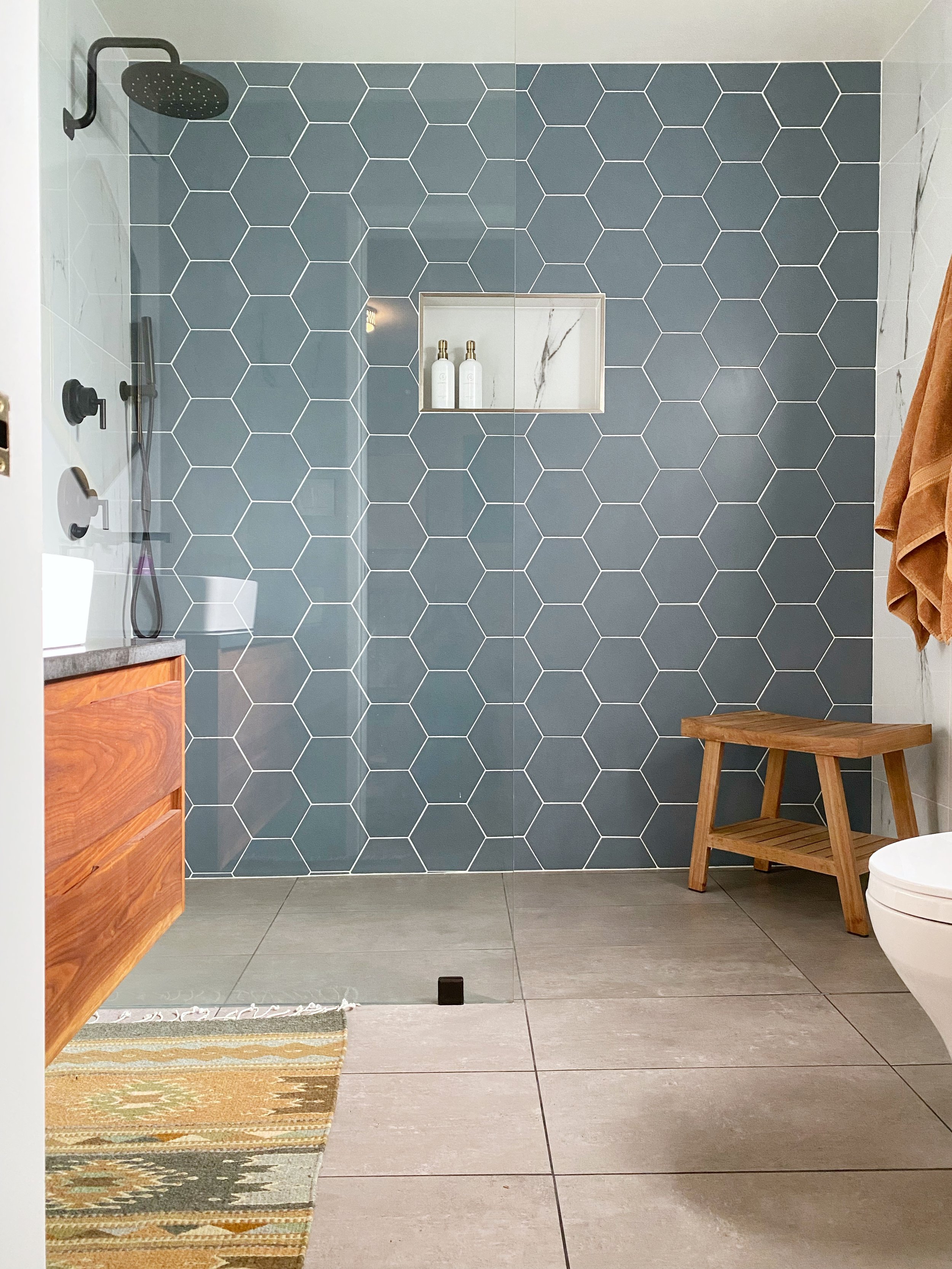 Modern bathroom with a walk-in shower featuring hexagon blue tiles, a built-in niche with two bottles, a wooden bench, a towel hanging on the wall, a partial view of a toilet, and a small striped rug on the floor.