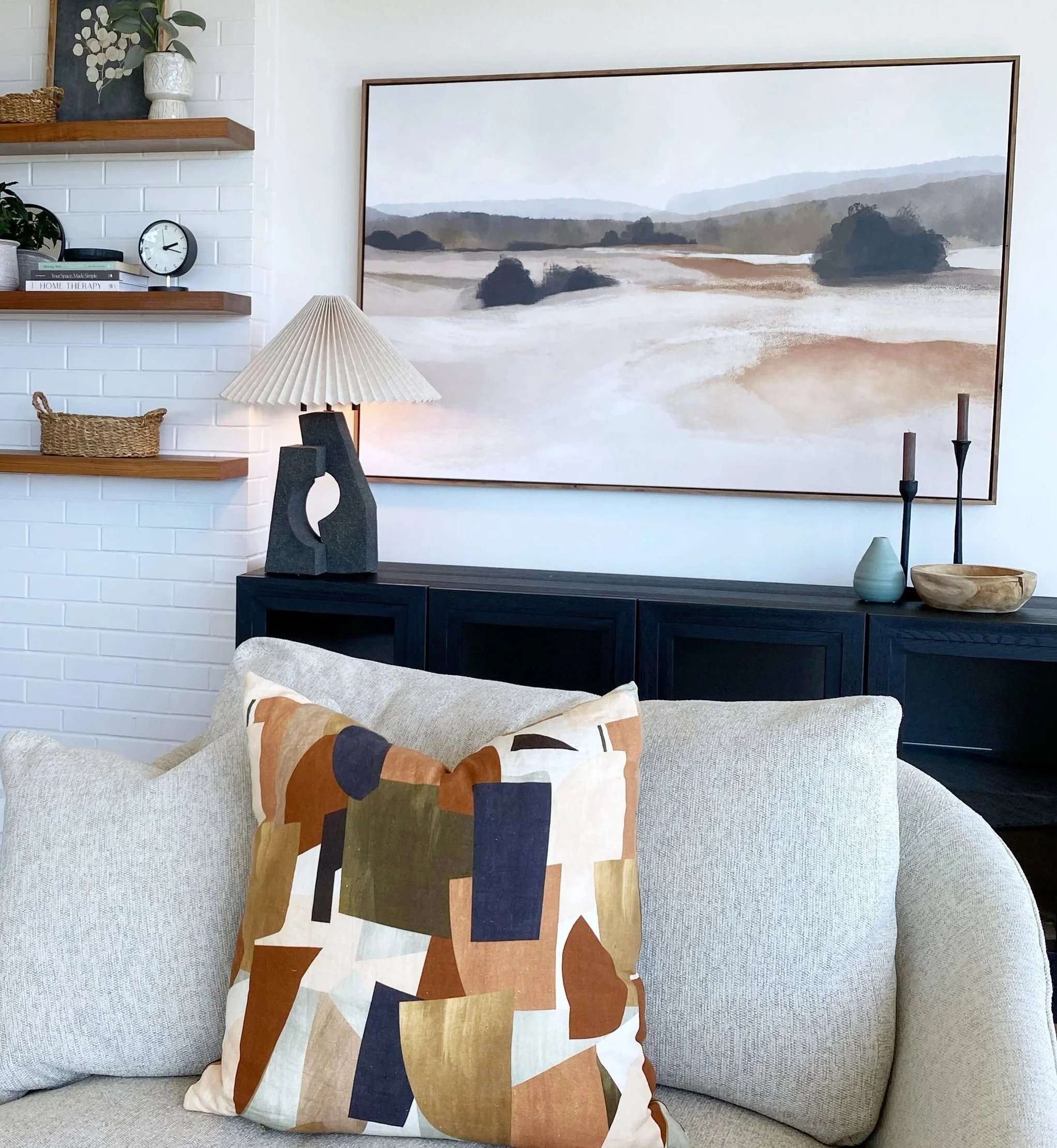 Living room with beige sofa, decorative pillow with abstract geometric pattern, black cabinet, large landscape painting, table lamp, and wall shelves with books, clock, and decorative baskets.
