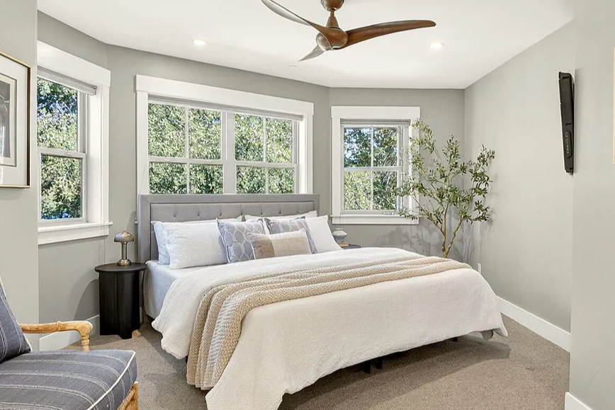 Bright bedroom with large windows, a gray upholstered bed with white and beige bedding, a black nightstand, a small lamp, an armchair with striped fabric, a ceiling fan, and a potted plant.