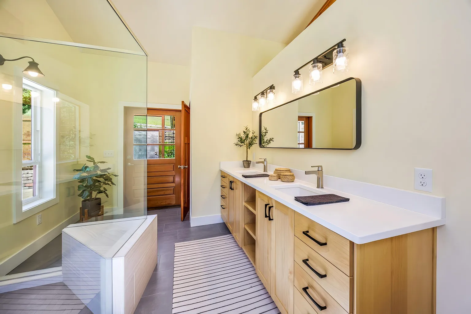 Modern bathroom with double vanity, large mirror, and glass-enclosed shower with a mirror reflection of a window and potted plant