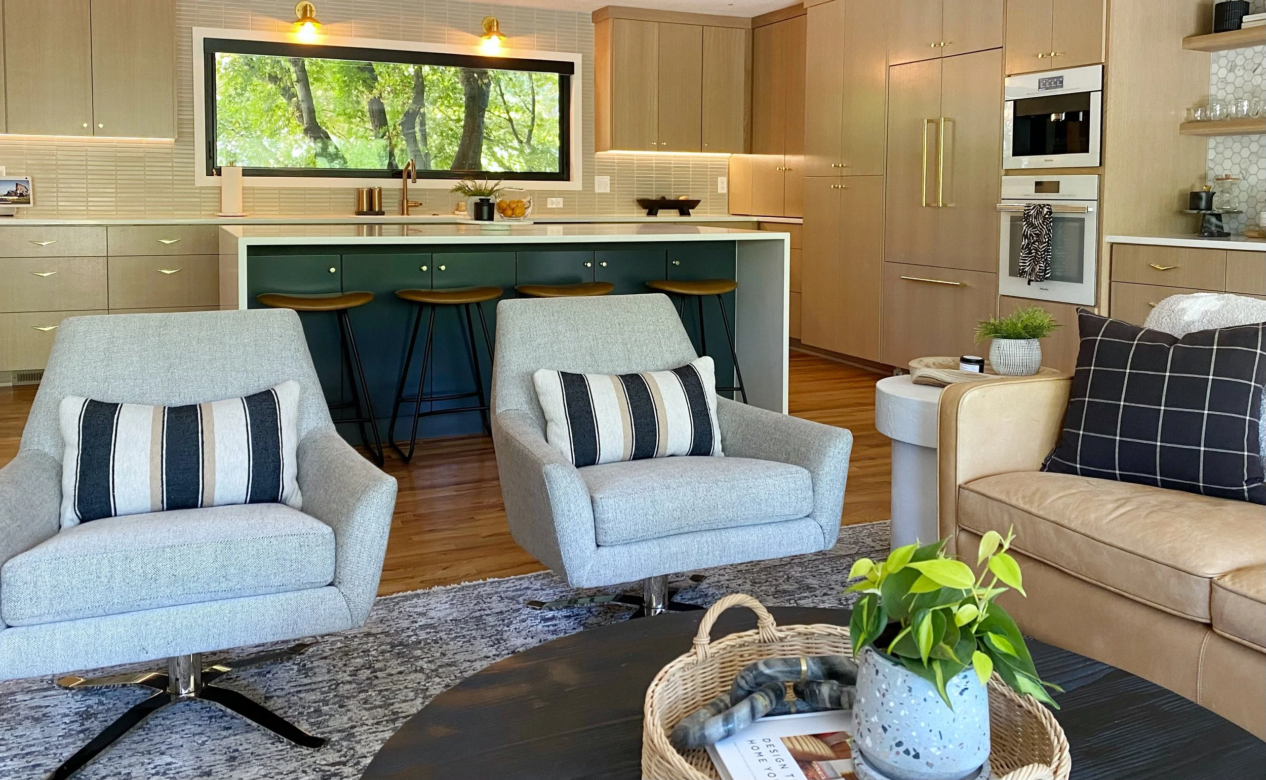 Living room with two light gray armchairs with striped pillows, a beige sofa with a black plaid pillow, a round black coffee table with a potted plant, and a kitchen in the background featuring a large window, a green island with barstools, and woode