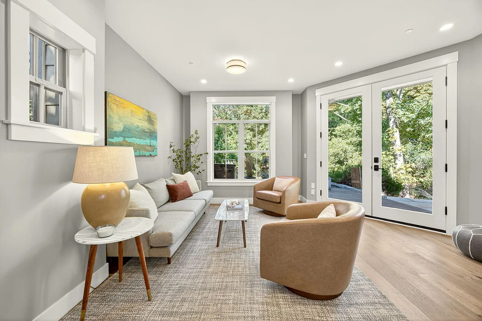 Bright living room with large windows and glass sliding doors, furnished with a beige sofa, two armchairs, a wooden coffee table, and decorative plants, overlooking a lush green outdoor area.