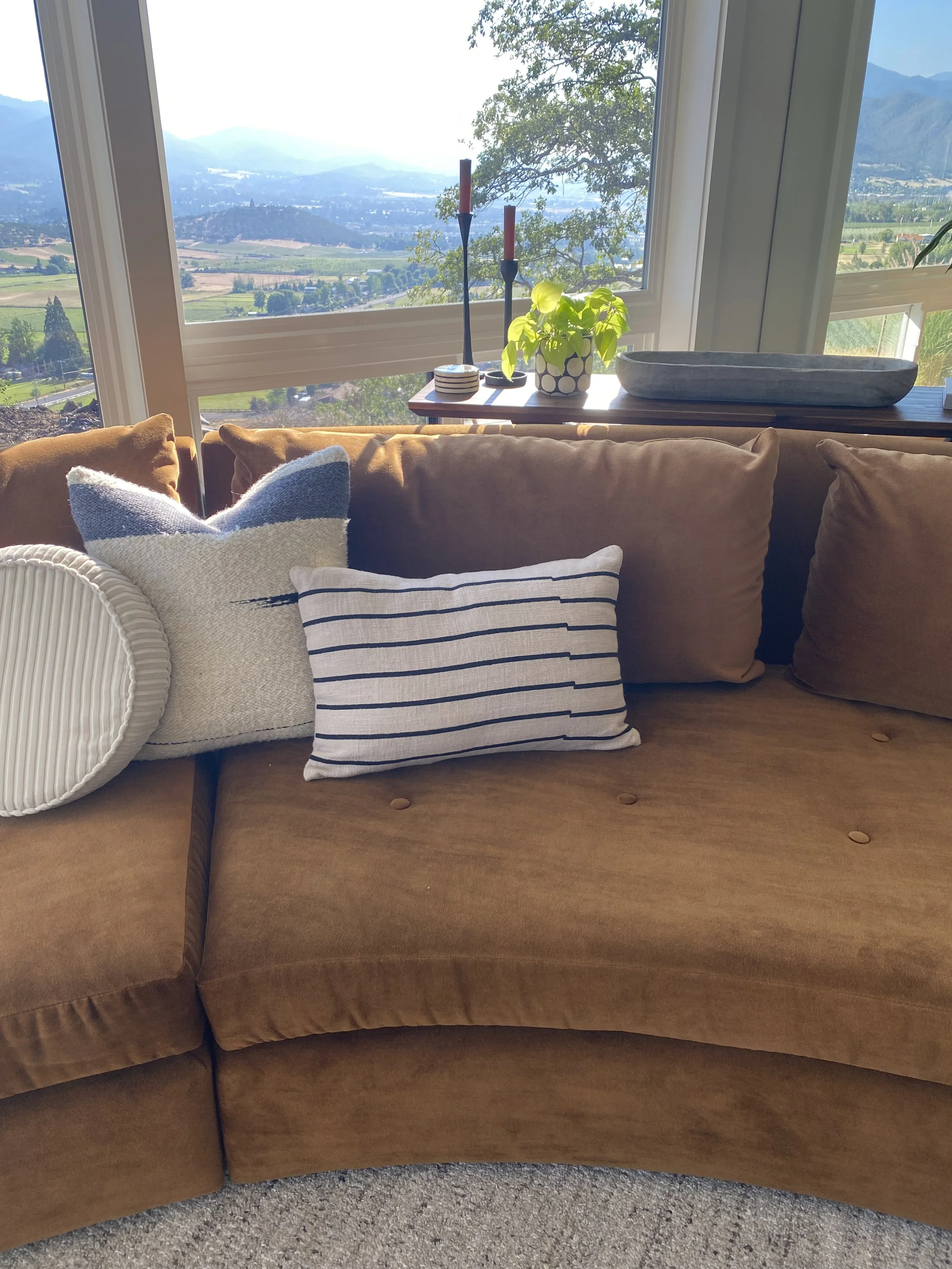 Living room with a brown couch, decorative pillows, and a view of green hills and mountains through large windows, with sunlight streaming in.