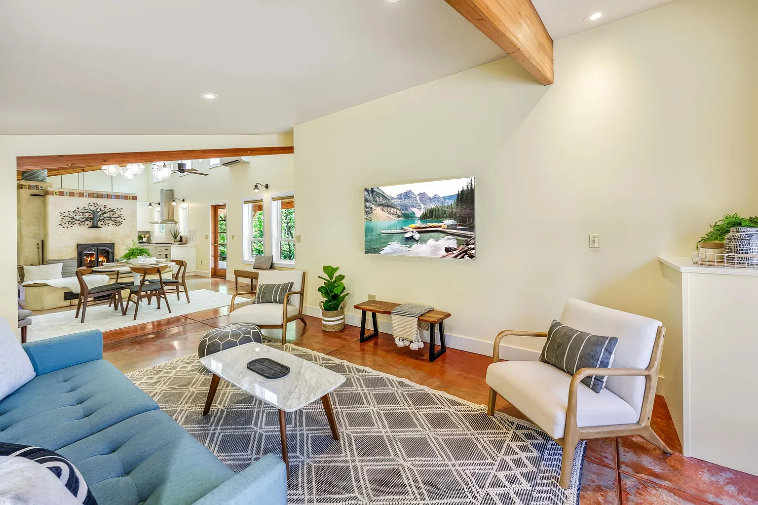 Living room with a blue sofa, two white armchairs, a marble coffee table, and a wall-mounted flat-screen TV showing a mountain landscape and lake scene. Nearby are a potted plant, a wooden bench, and pillows on the chairs. The room features hardwood 