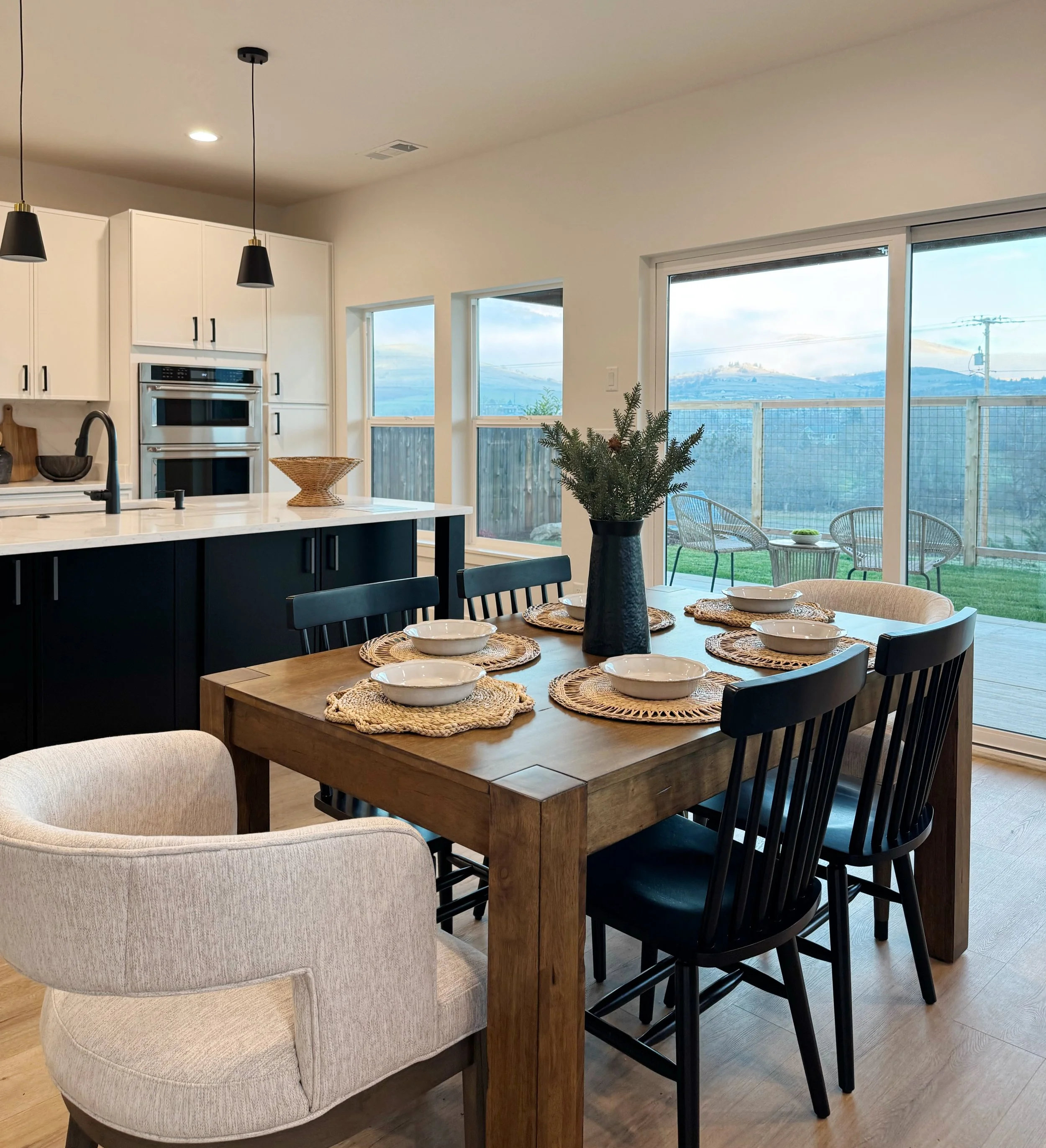 Dining area with a wooden table set with plates, bowls, woven placemats, and a black vase with greenery, adjacent to a sliding glass door leading to an outdoor patio with chairs and a grassy yard, connected to a white and black kitchen.