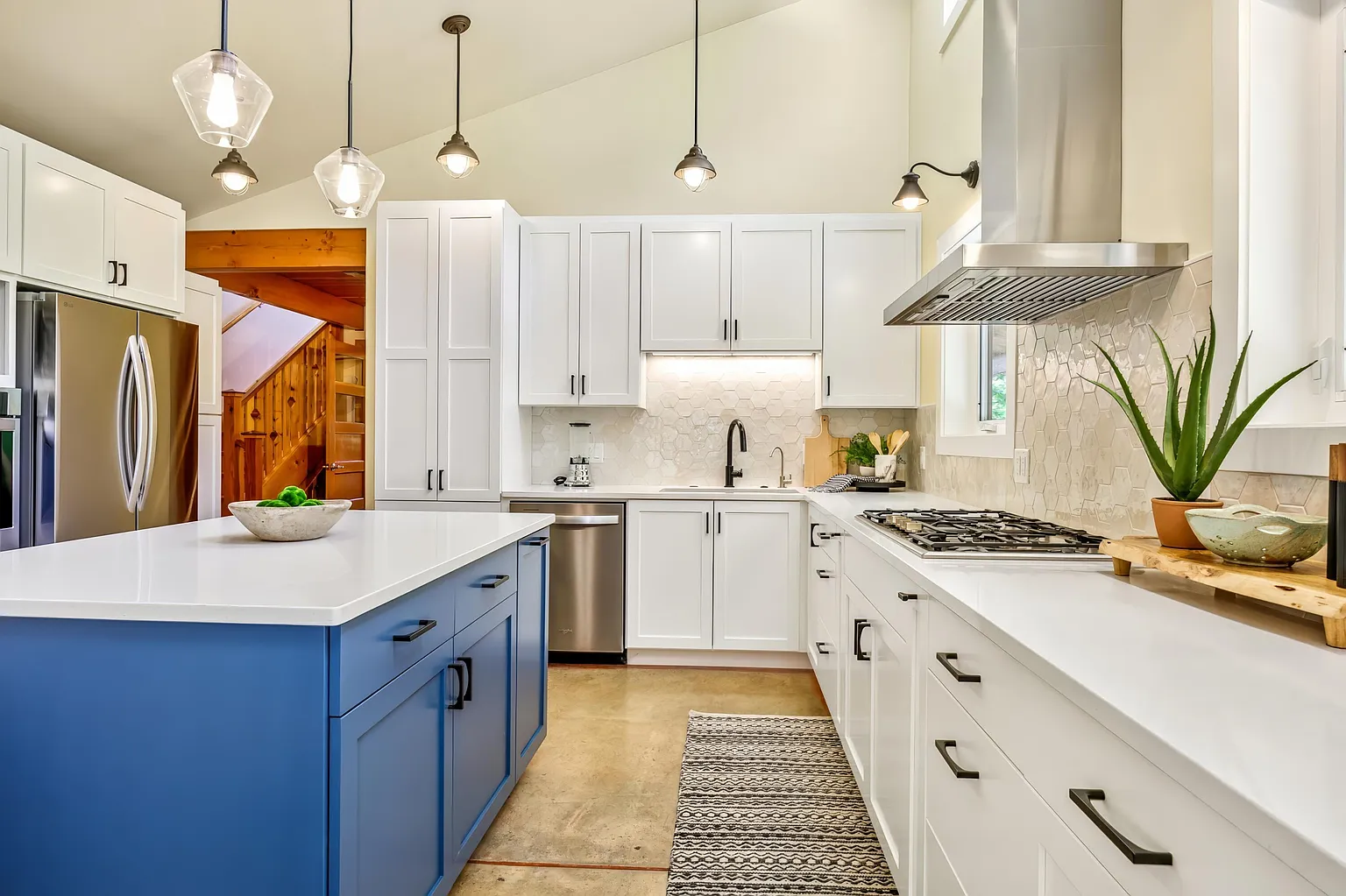 Modern kitchen with white cabinets, an island with a blue base, stainless steel refrigerator, and a gas stove. Decor includes a potted aloe vera plant and a woven rug.