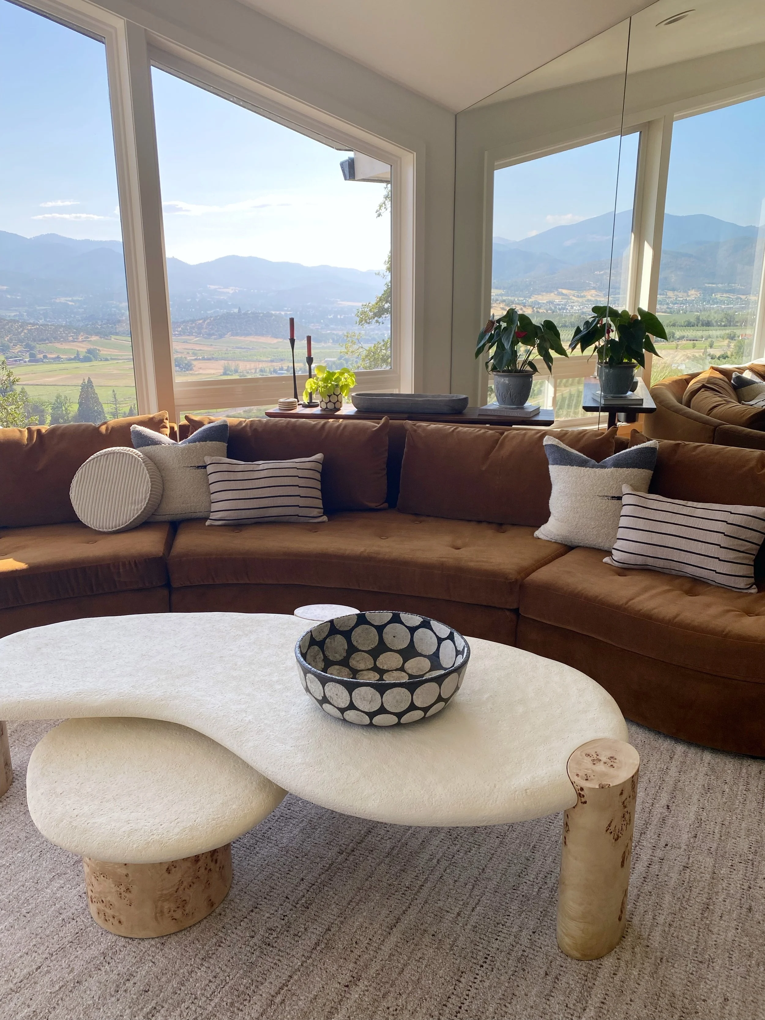 A living room with a large brown sectional sofa decorated with striped and solid throw pillows, a modern cream-colored coffee table with a rounded organic shape and wooden legs, and large windows showing a scenic view of mountains, fields, and a clea