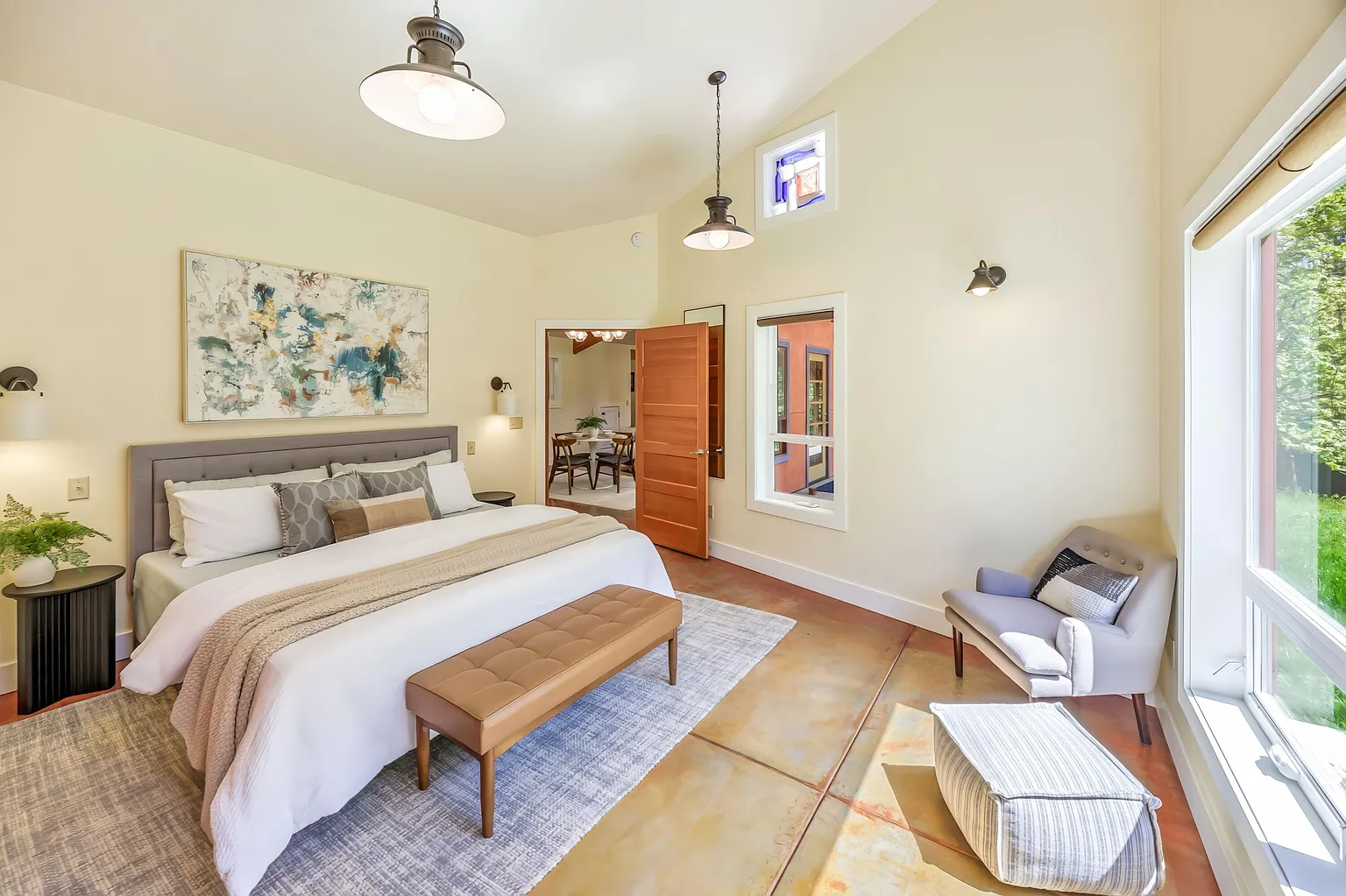 Bright bedroom with a large bed, artwork above the headboard, a bench at the foot of the bed, side tables with lamps, a gray armchair, and large windows showing greenery outside.