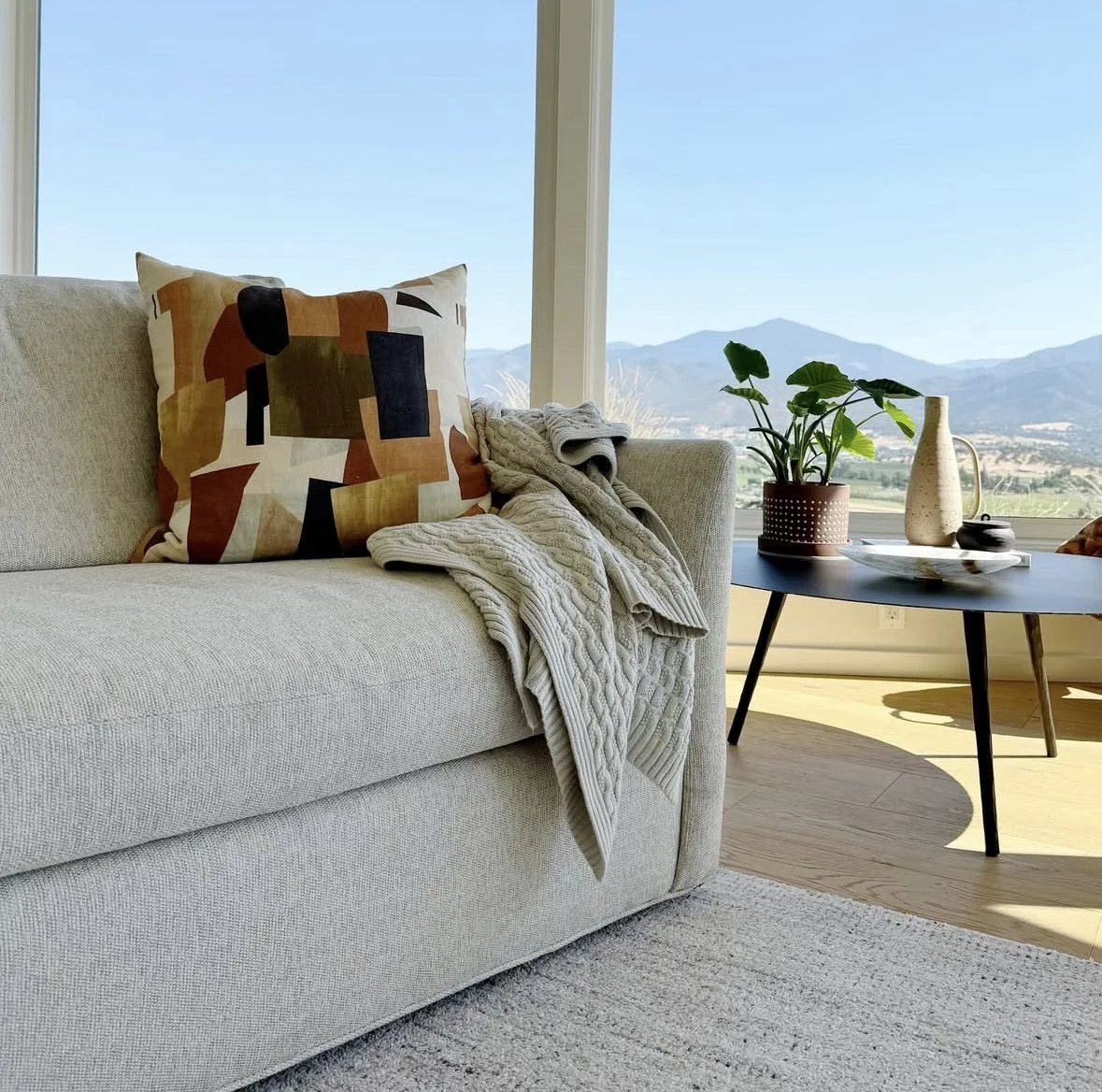 Living room with a cream-colored sofa, abstract decorative pillow, gray textured blanket, side table with potted plant, ceramic pitcher, and bowl, large window with mountain view.