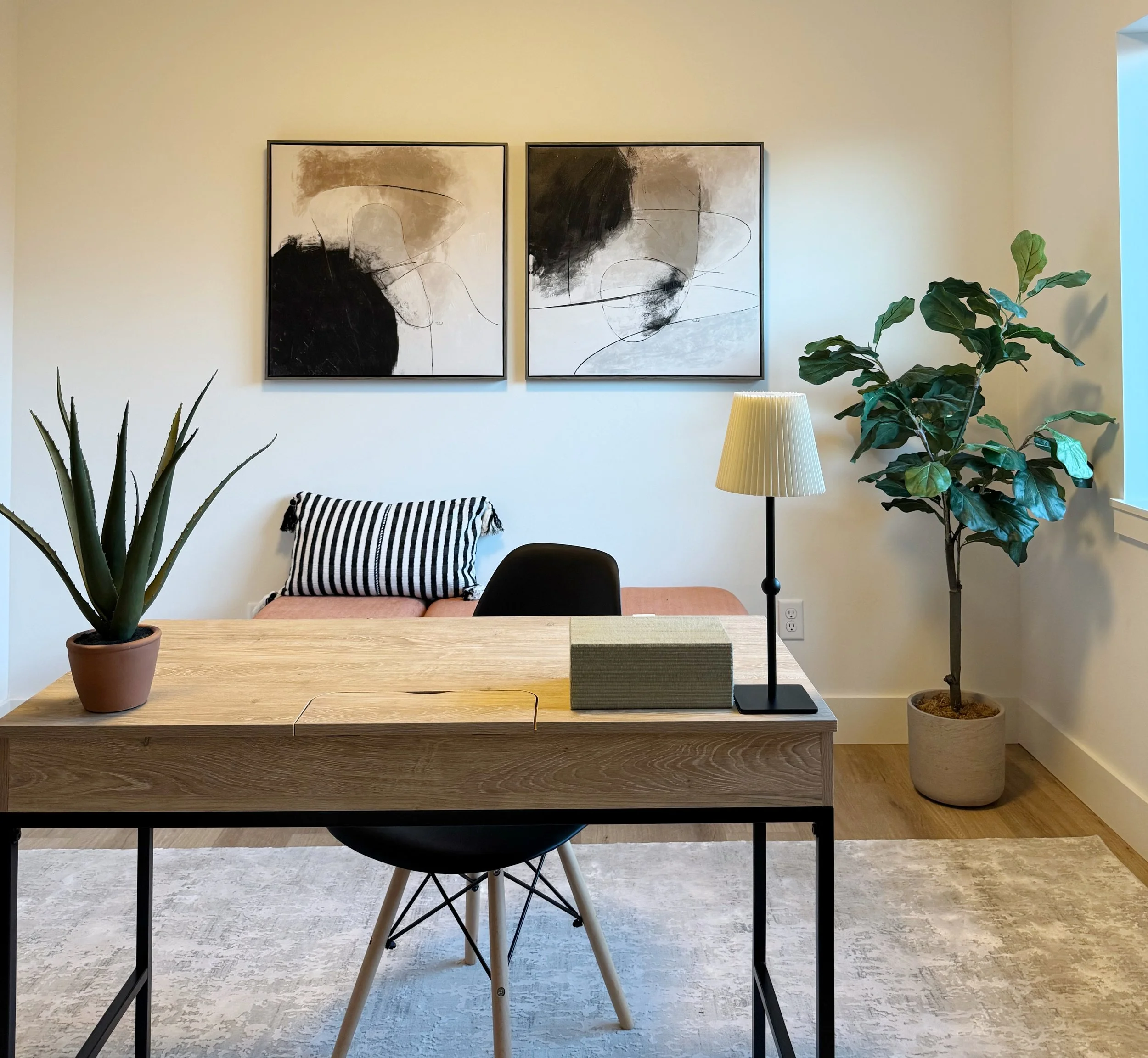 Modern office or living room with a wooden desk, potted aloe vera plant, table lamp, abstract black and white artworks, black chair, pink cushioned bench with striped pillow, and large potted fiddle leaf fig plant near the window.
