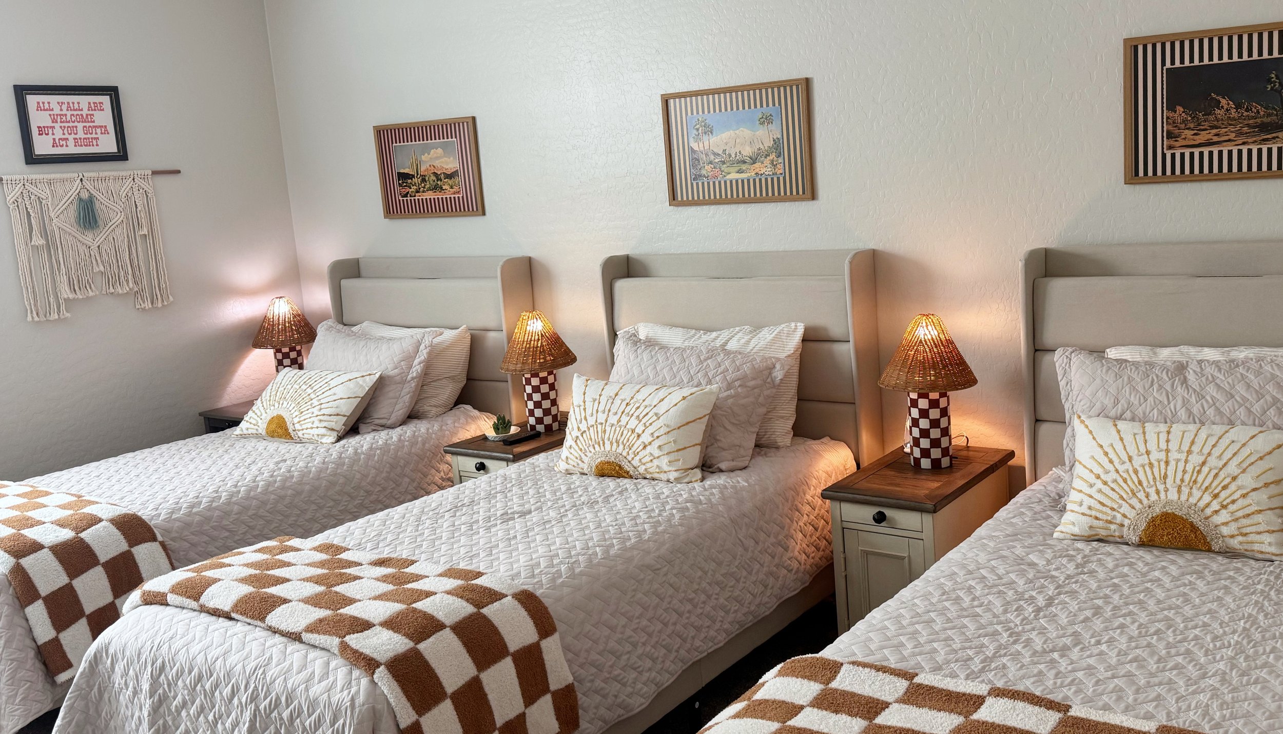 Three guest beds with white quilted bedding, decorative pillows with sunburst patterns, each bed has a bedside table with a checkered lamp, and framed art and wall decor above.