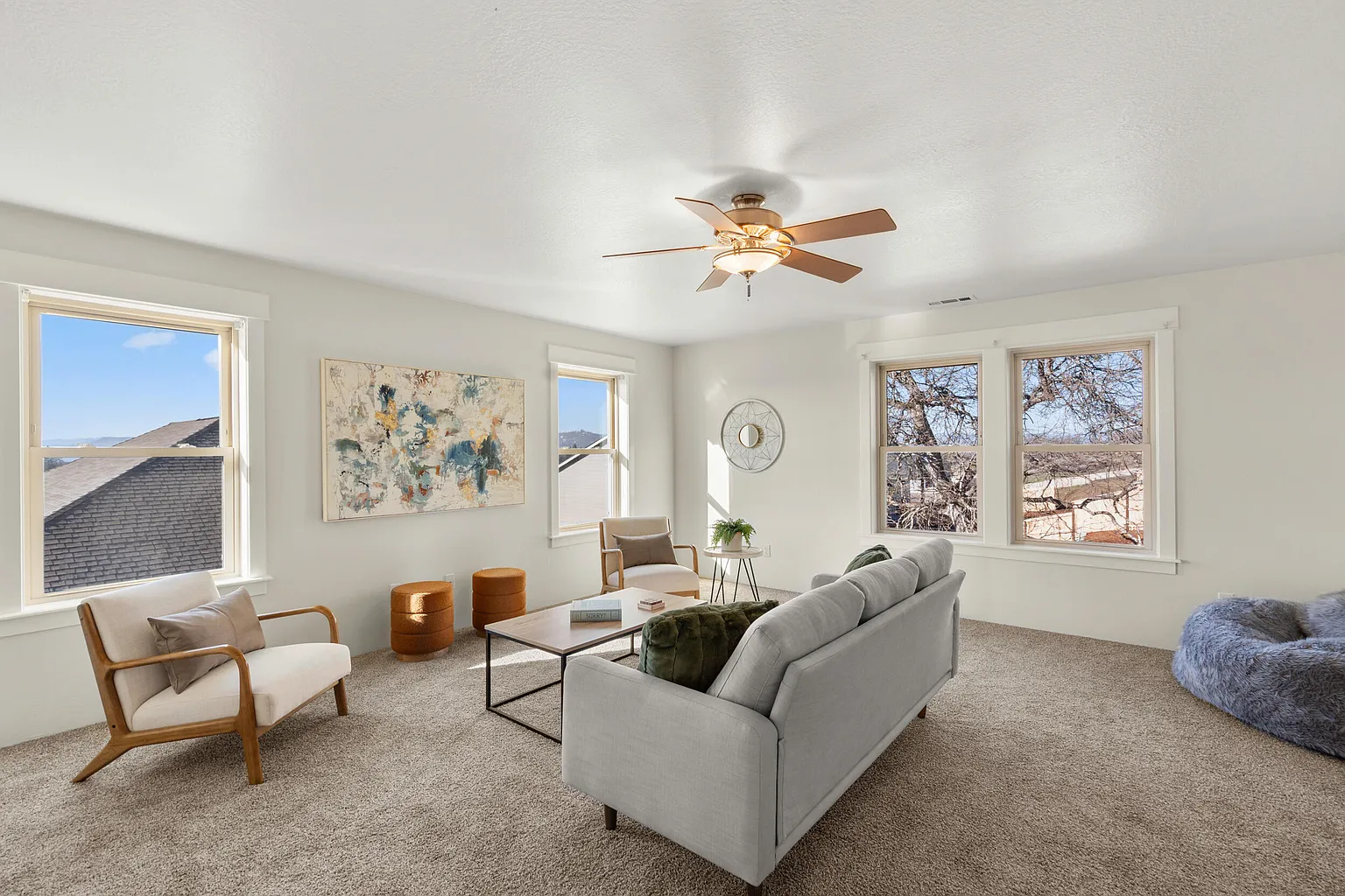 Bright living room with three windows, a ceiling fan, a light gray sofa, a beige armchair, a white side table with a plant, two brown ottomans, a wooden armchair, a textured bean bag, a colorful abstract wall art, and a circular mirror.