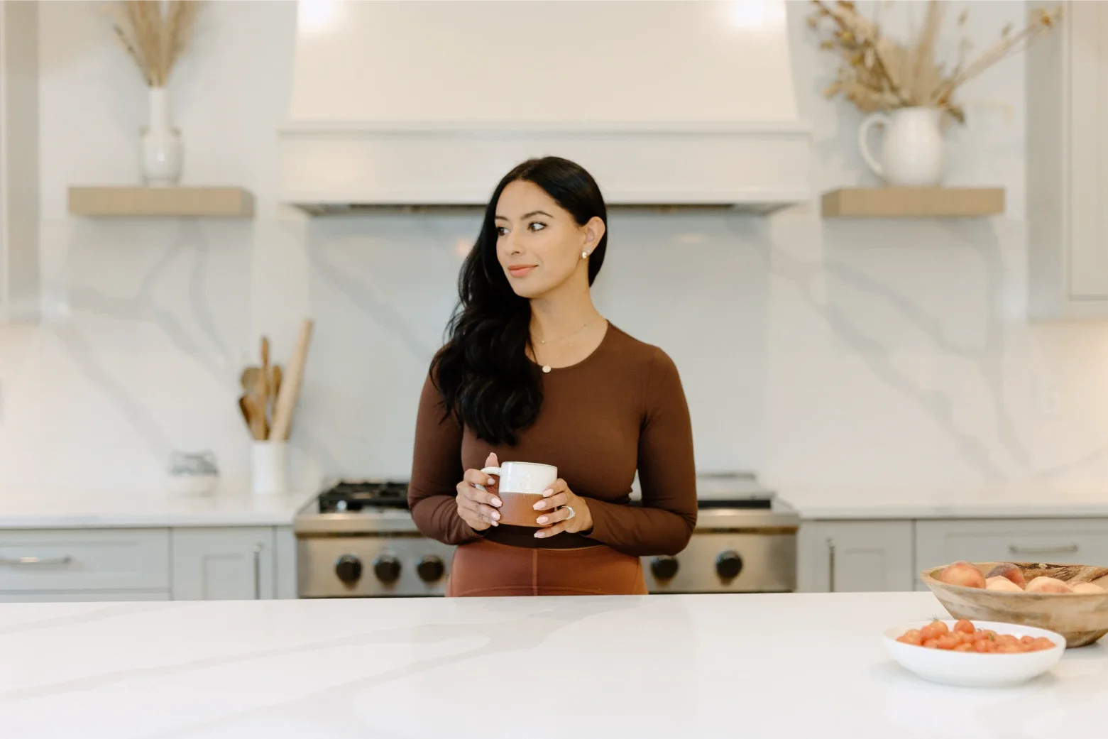 A woman with long black hair and a brown dress holding a mug in a modern kitchen with white cabinets and countertops, and bowls of fruit on the counter.