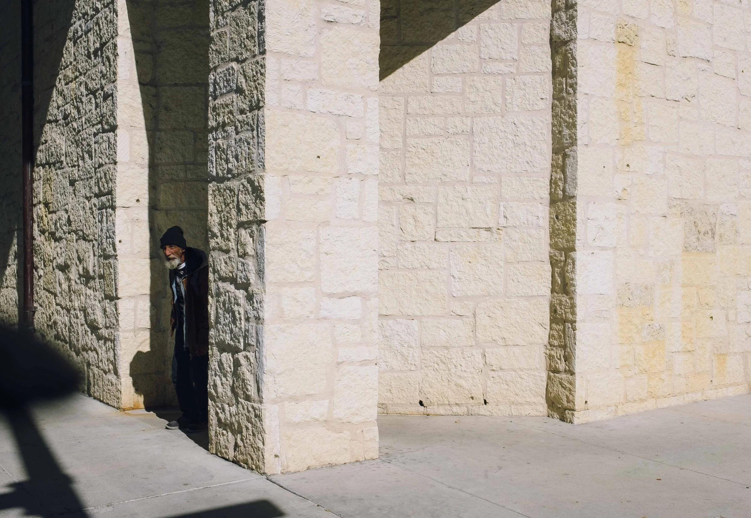 An elderly man with a black beanie, beard, and dark clothing peeks out from behind a stone wall corner, standing on a paved sidewalk near a large stone building.