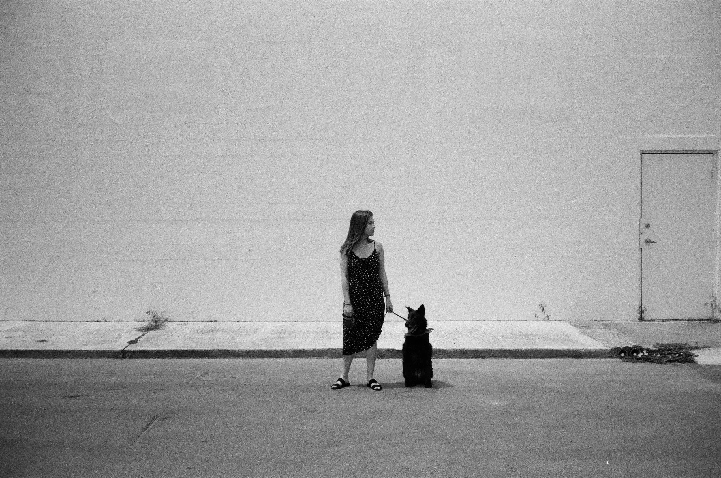 A woman standing on a sidewalk holding a dog on a leash in front of a plain wall with a door, black and white photo.