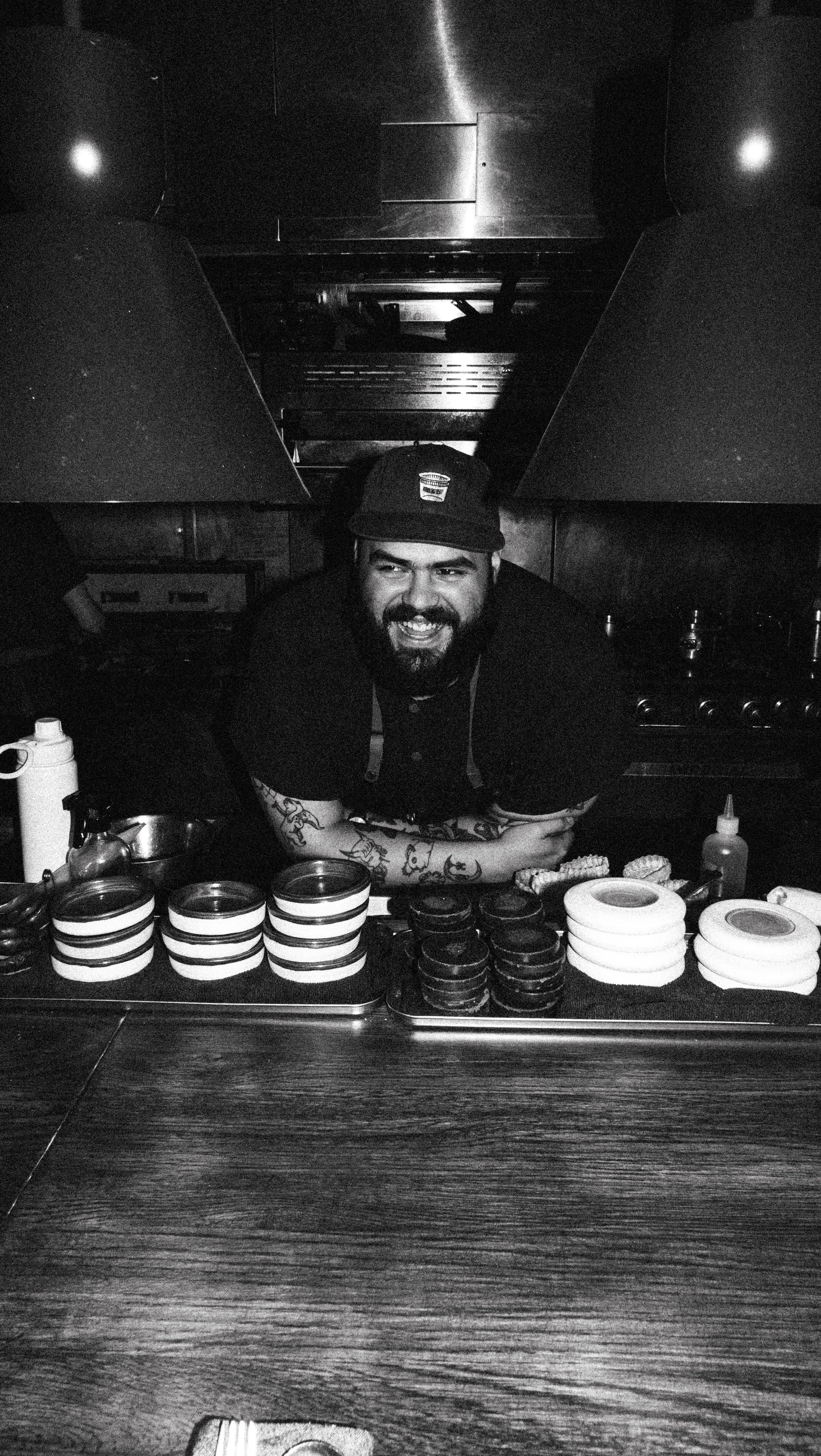 A man with a beard and tattoos smiling behind a counter with various sushi rolls and bowls, in a dimly lit kitchen or sushi bar environment.