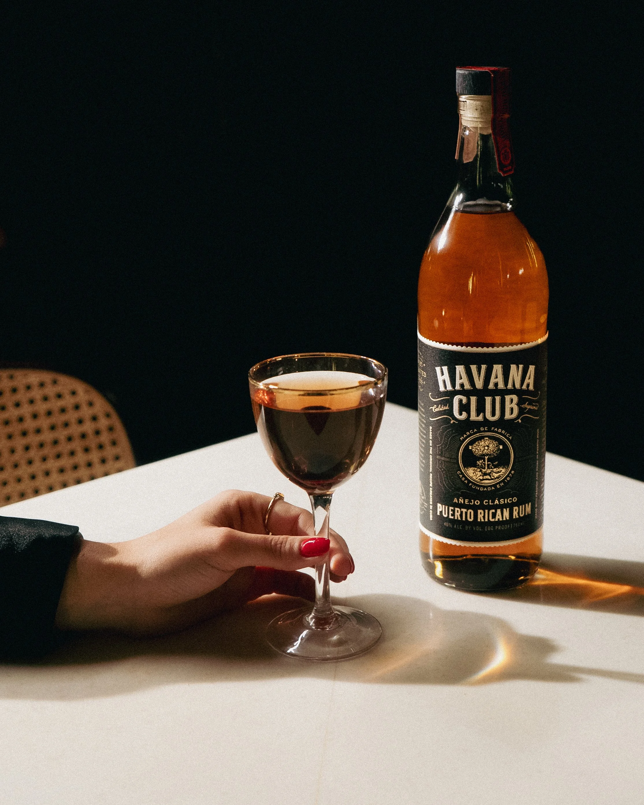 A hand with red nail polish holding a glass of dark amber liquid next to a bottle of Havana Club Puerto Rican Rum on a white table.