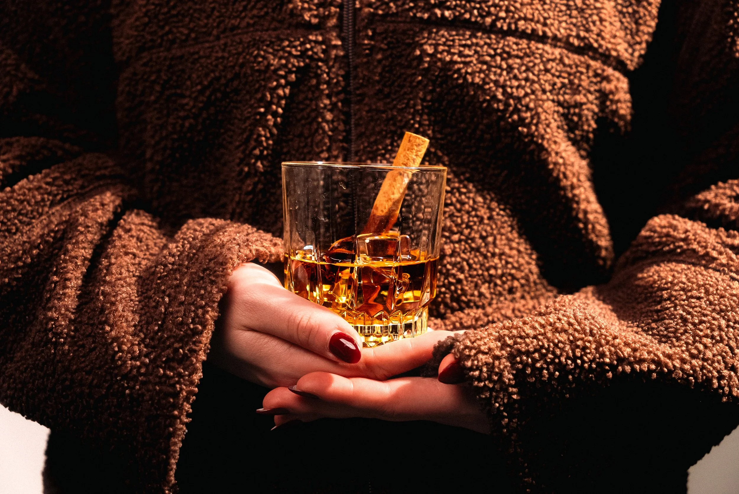 Person holding a glass of whiskey with ice and a cinnamon stick, wearing a brown textured fleece jacket.