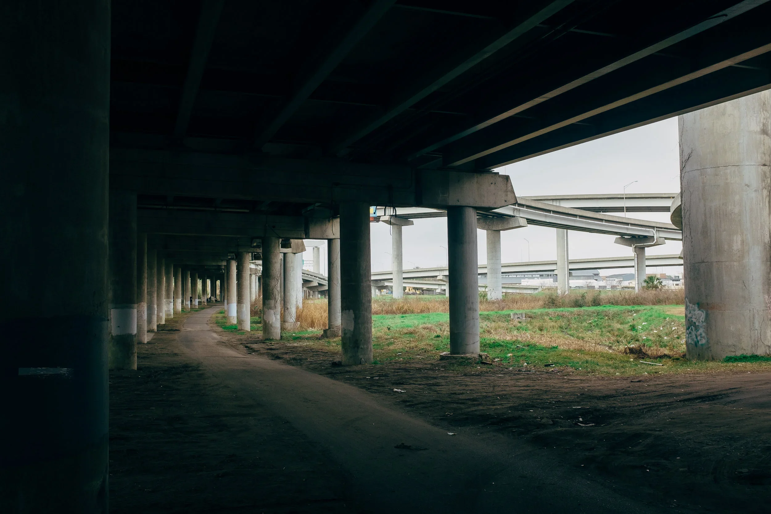 Underground highway overpass with concrete pillars and a dirt path underneath, with grassy areas and additional highway structures visible in the background.