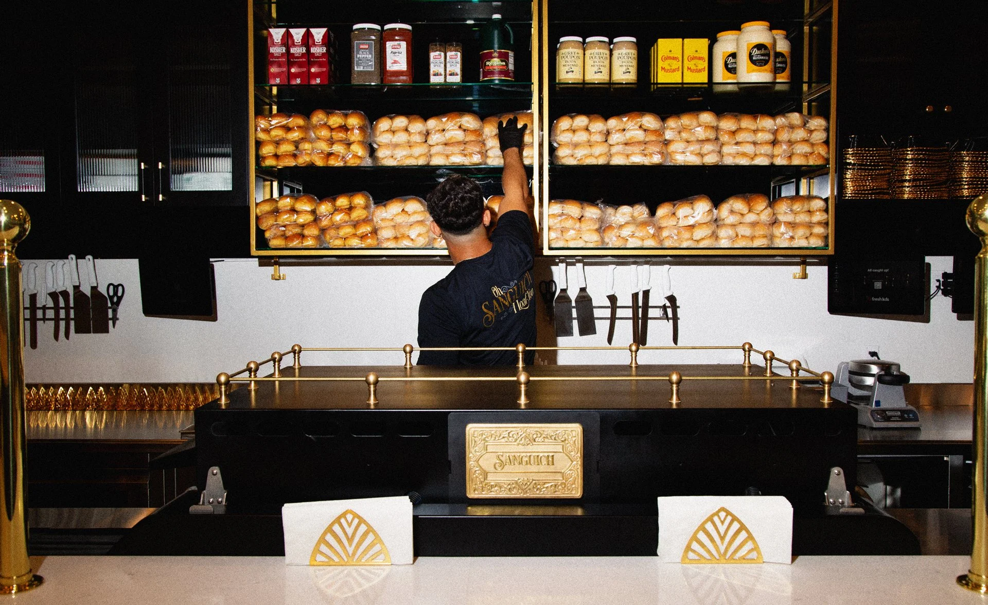 Worker stocking shelves with buns and condiments in a deli or burger shop.