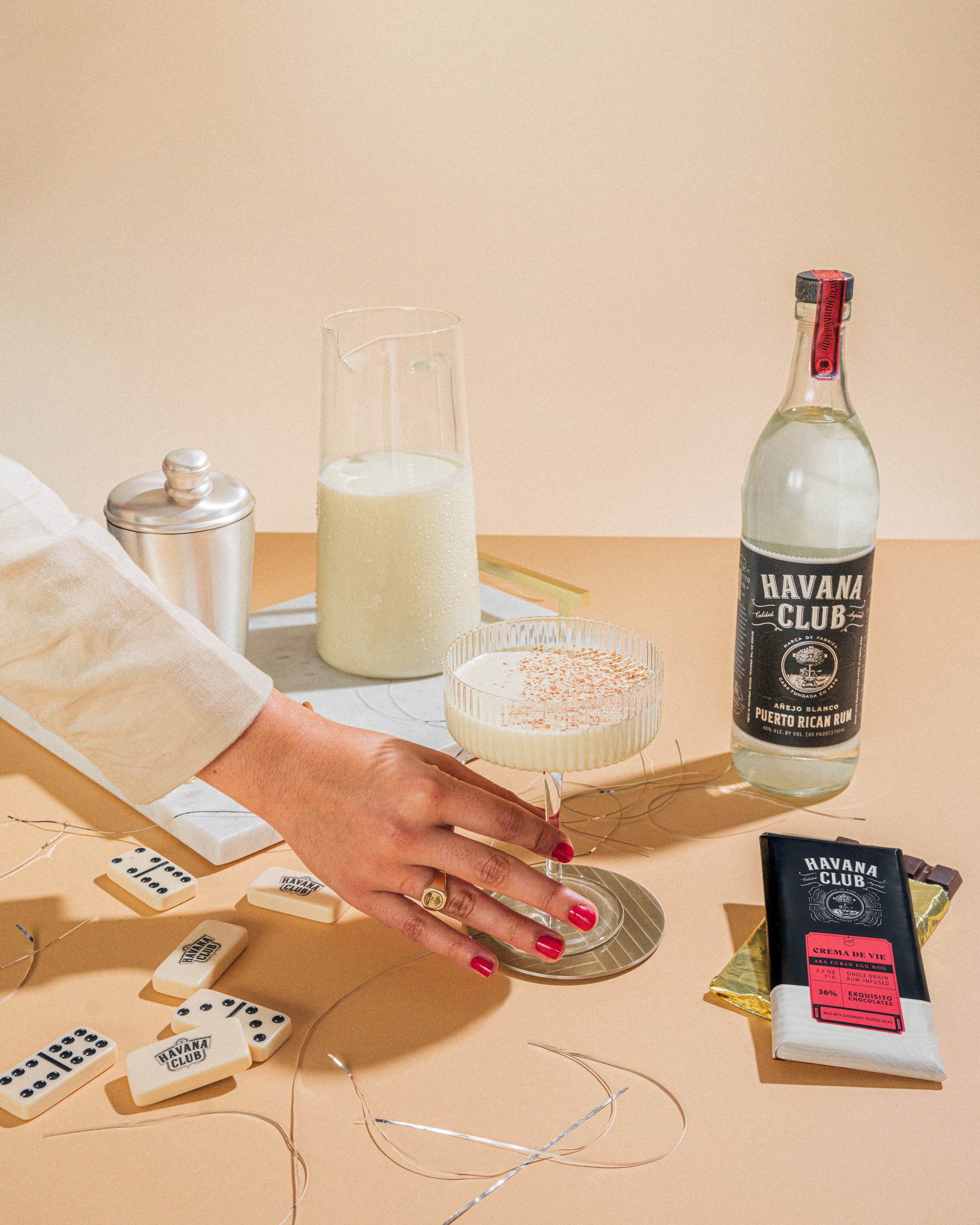 Woman's hand with red nail polish holding a cocktail glass on a beige table, surrounded by a bottle of Havana Club rum, a glass pitcher of milk, a container, domino tiles, a chocolate bar, and decorative string lights.