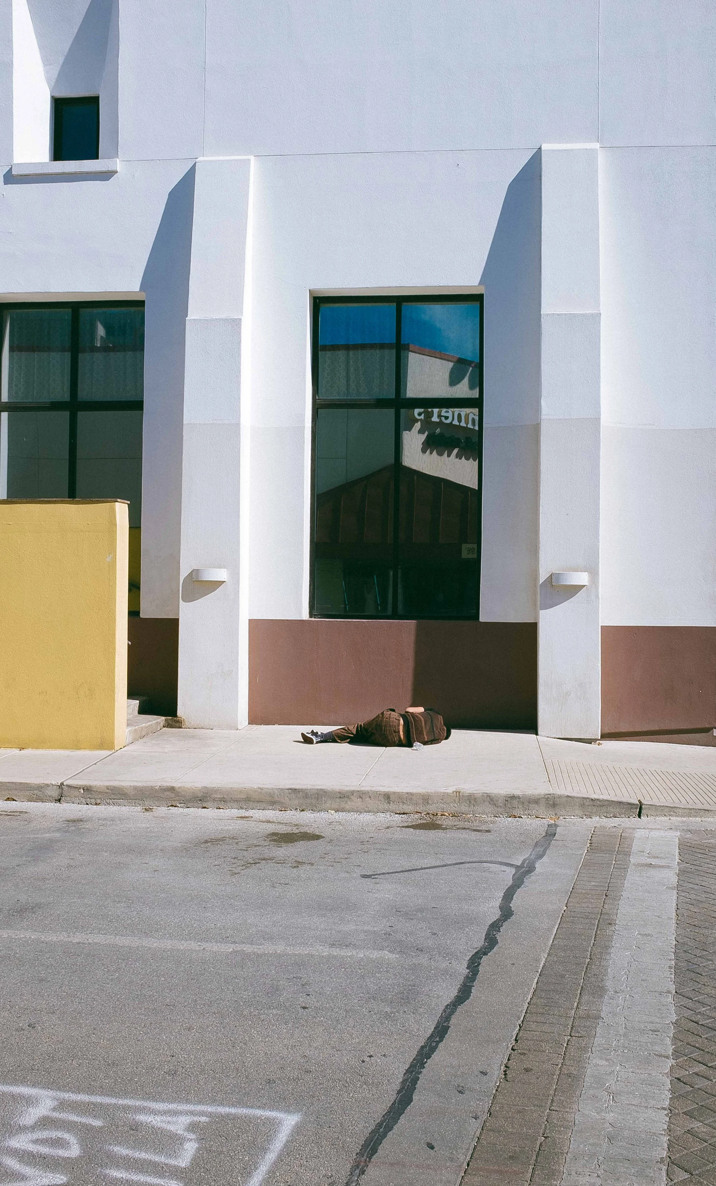 A person lying on the sidewalk near a white building with large windows and a yellow wall.