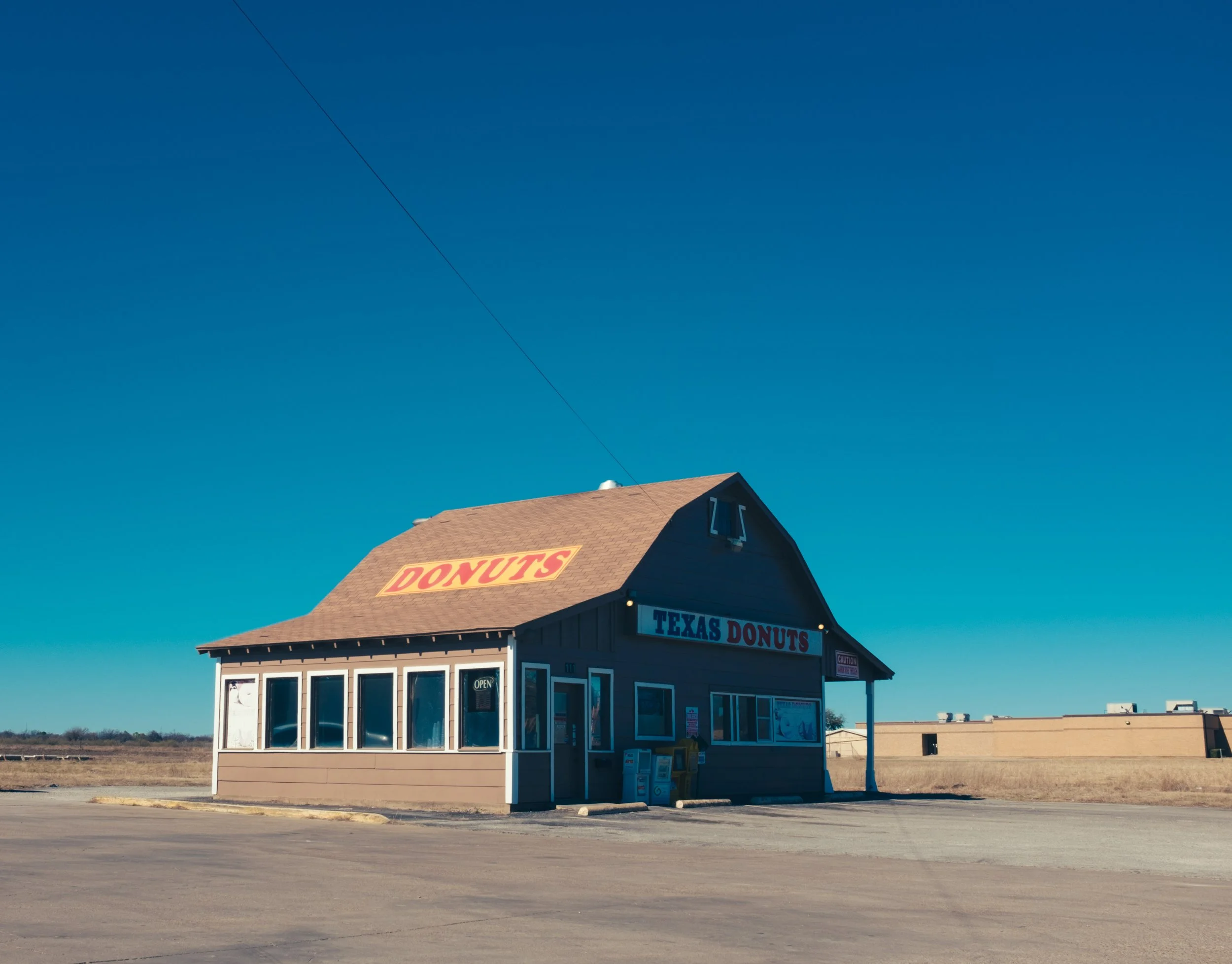 Small, brown roadside building with a sign that says 'Donuts' on the roof and 'Texas Donuts' on the front, surrounded by a mostly empty parking lot under a clear blue sky.