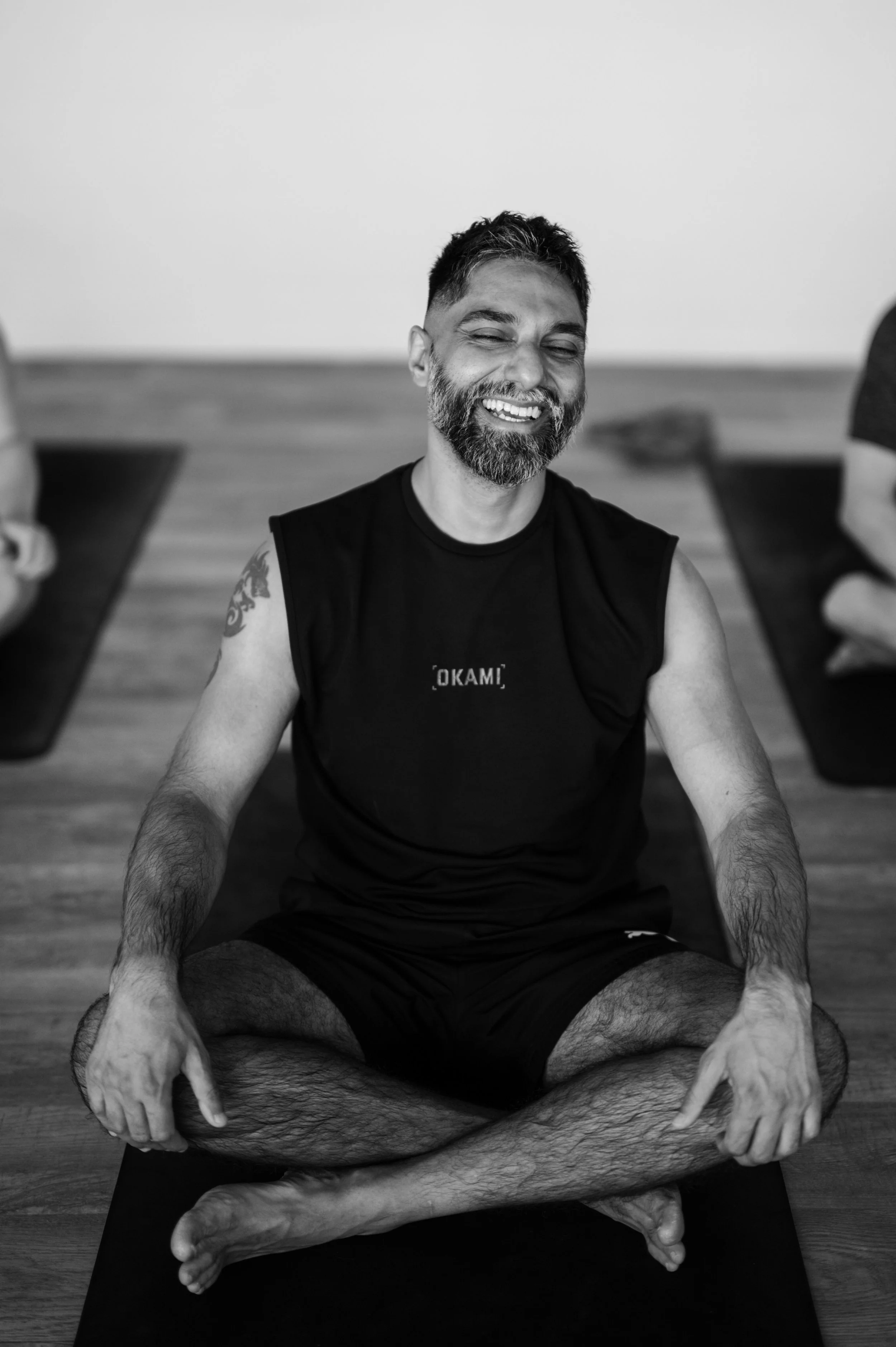A man with a beard sitting cross-legged on a yoga mat, practicing yoga or meditation indoors, with a smiling expression.