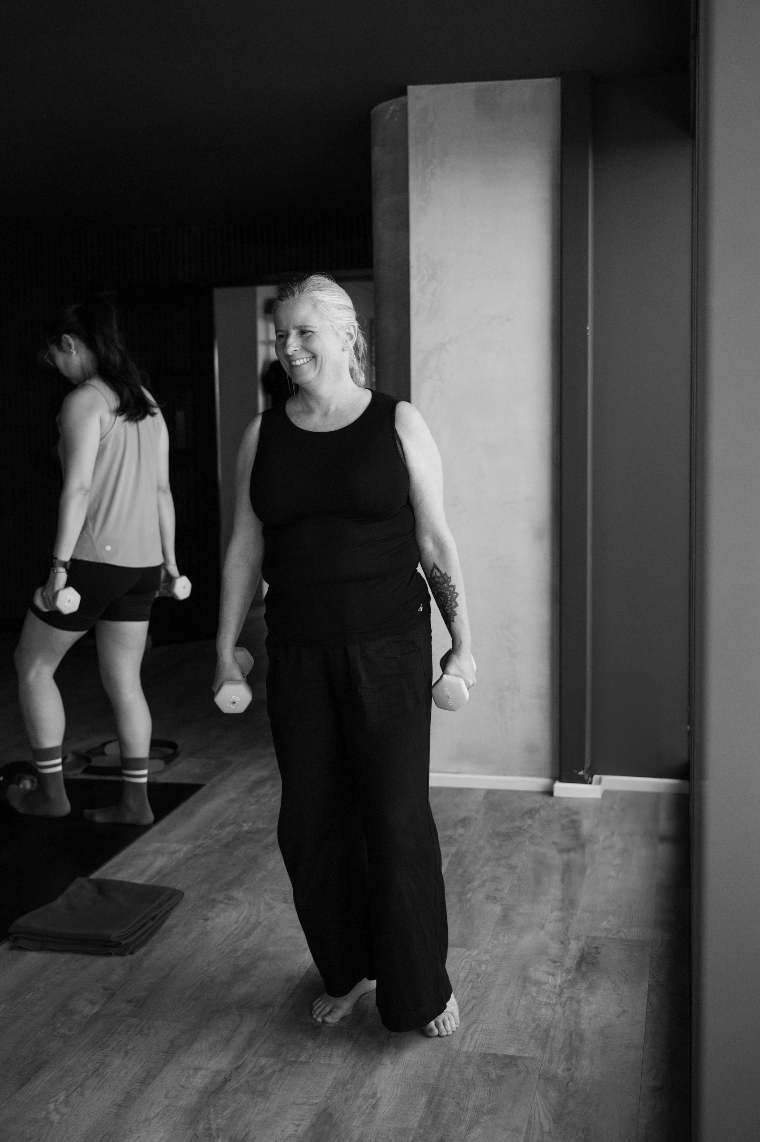 A woman with gray hair smiling and holding dumbbells in a workout studio, with another person in workout attire in the background.