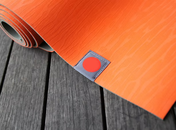 Close-up of an orange yoga mat with a circular red sticker on it, placed on a wooden deck.