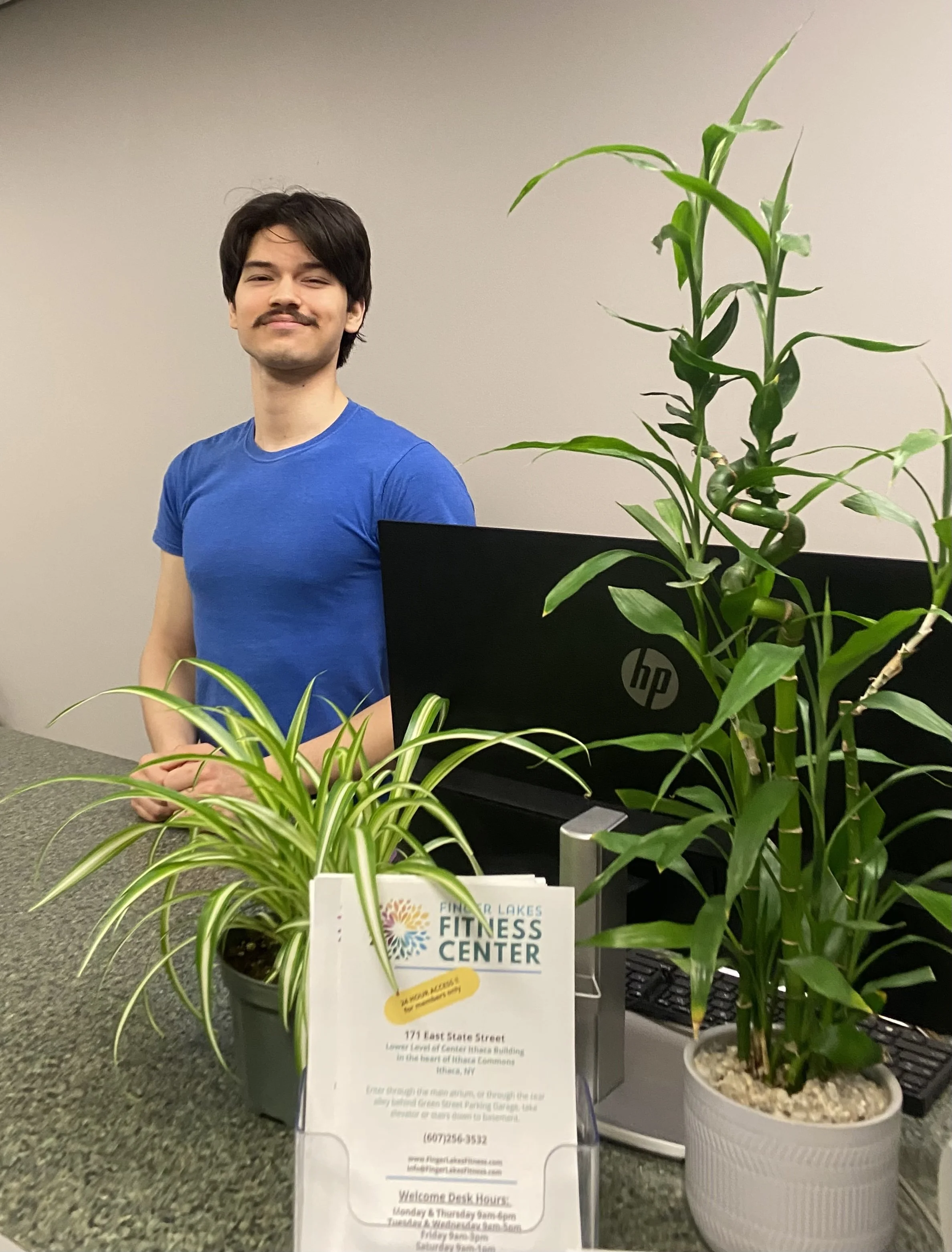 Front desk person standing behind the Welcome Desk that has potted plants on it along with a Finger Lakes Fitness Center sign.