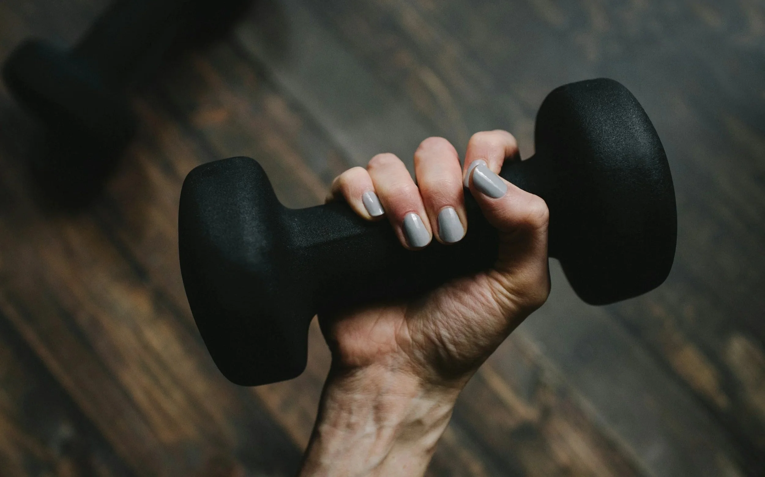 Close-up of a person's hand holding a black dumbbell with gray-painted nails, on a wooden gym floor.